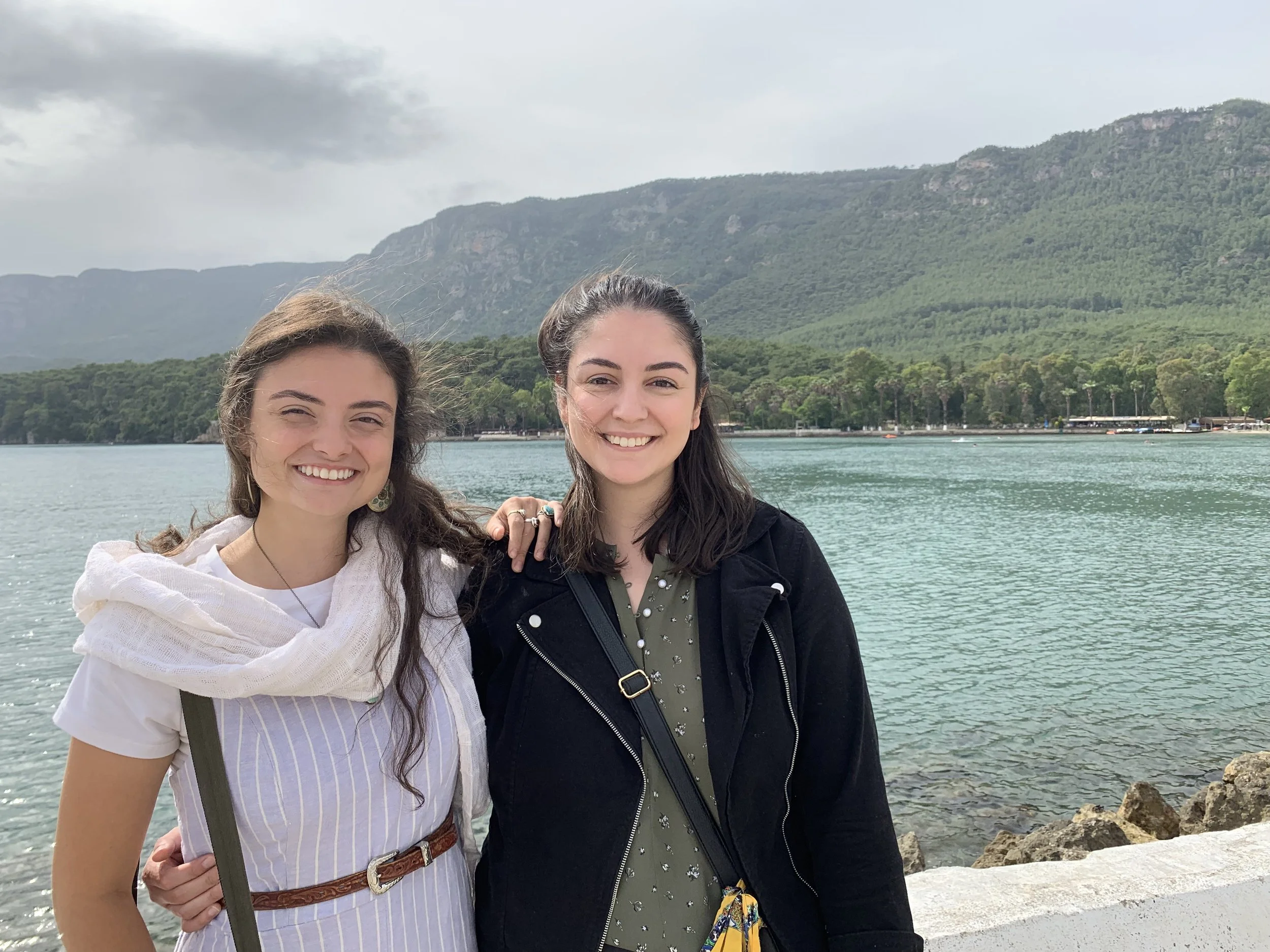Two young women smiling near water with mountains in the background.