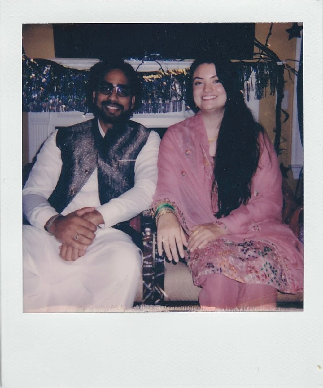 A man and woman sitting together at a celebration, dressed in traditional clothing, with festive decorations in the background.