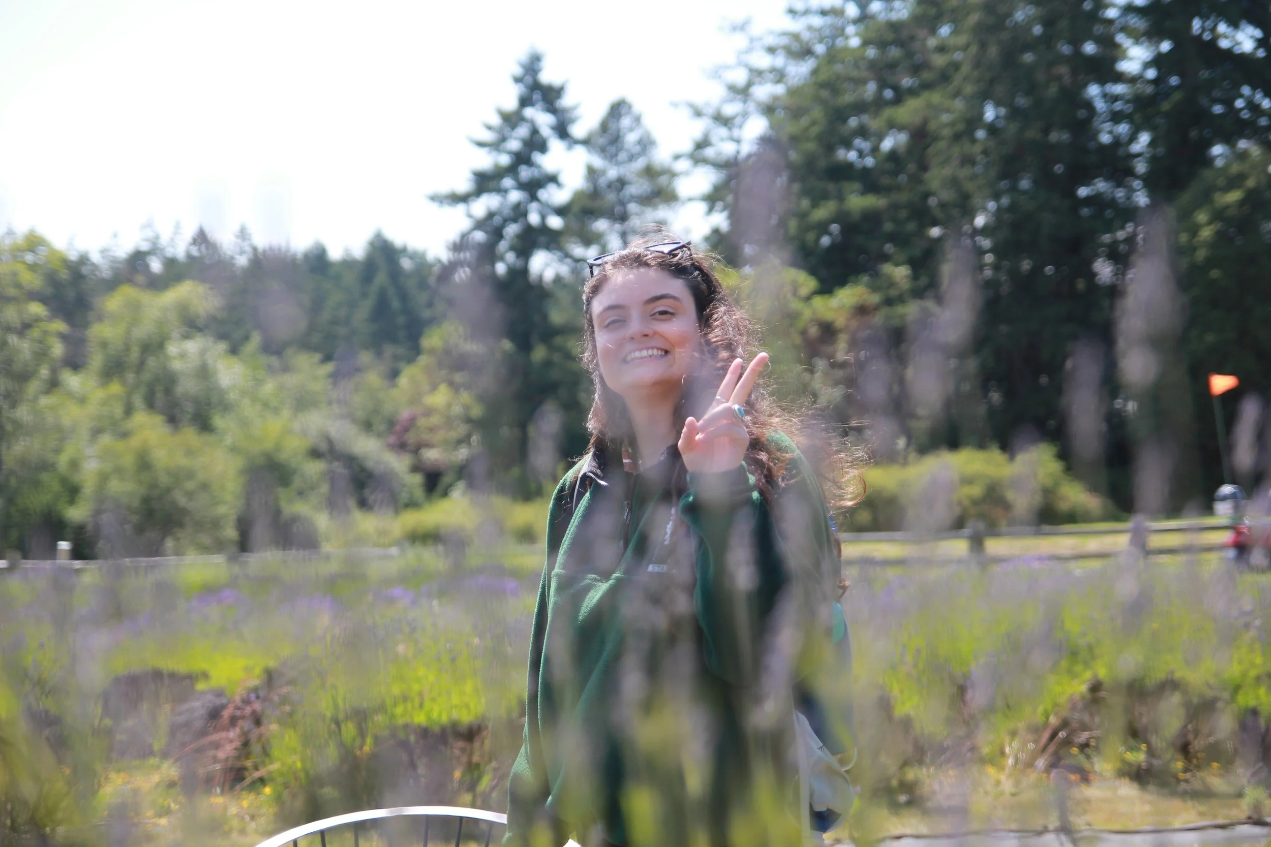 A smiling woman with curly hair making a peace sign with her fingers in front of a fence.