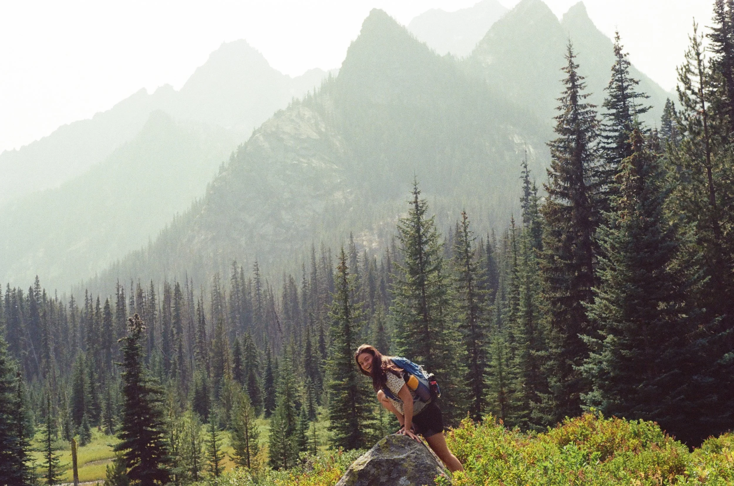 A woman in outdoor clothing posed with one knee on a large rock amid a forest of tall pine trees and mountainous landscape in the background.