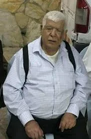 Older man with white hair wearing a light blue checkered shirt and black backpack, standing indoors near a stone wall.