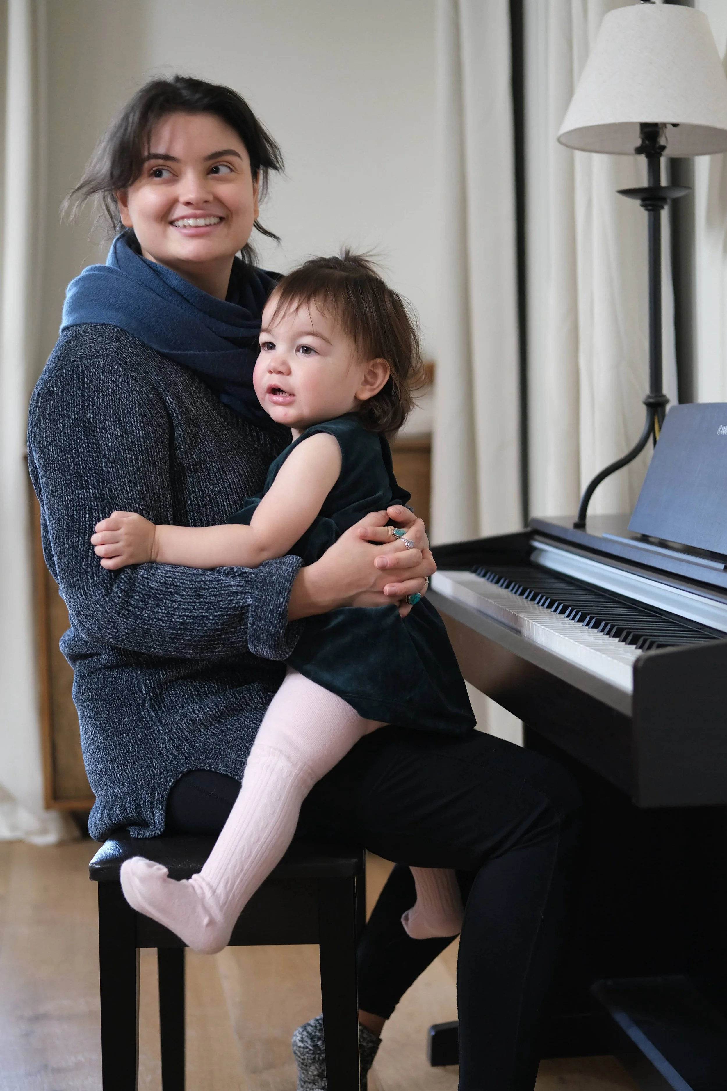 A woman holding a young girl on her lap while seated at a piano. The woman is smiling and looking off to the side. The girl is wearing a dark dress, pink tights, and patterned socks, with her arms around the woman’s neck. A floor lamp and curtains are in the background.