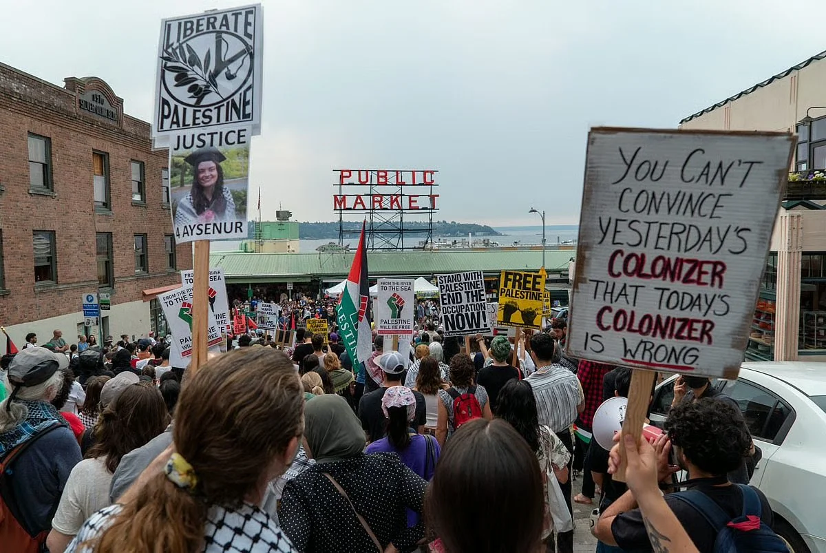 Crowd of protesters holding signs and banners in a demonstration supporting Palestine, with signs reading "Liberate Palestine Justice," and other messages about ending occupations and supporting Palestine.