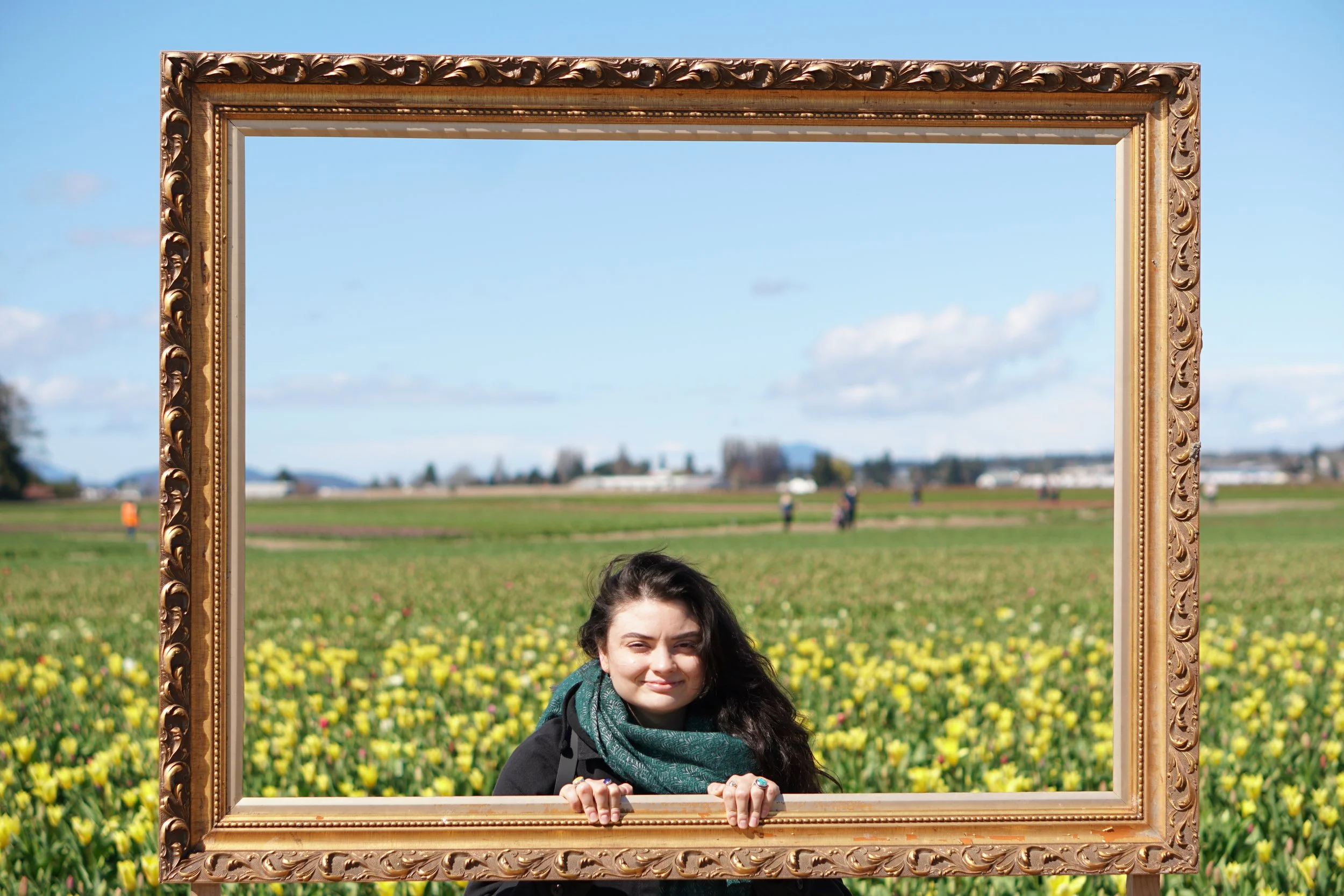 A woman with dark hair and a scarf around her neck poses behind a large ornate gold picture frame outdoors in a field of yellow and pink flowers with a blue sky and some clouds in the background.