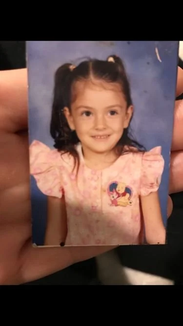 A young girl with dark hair in pigtails, wearing a pink dress with puffed sleeves and a Disney princess logo, smiling against a blue background.