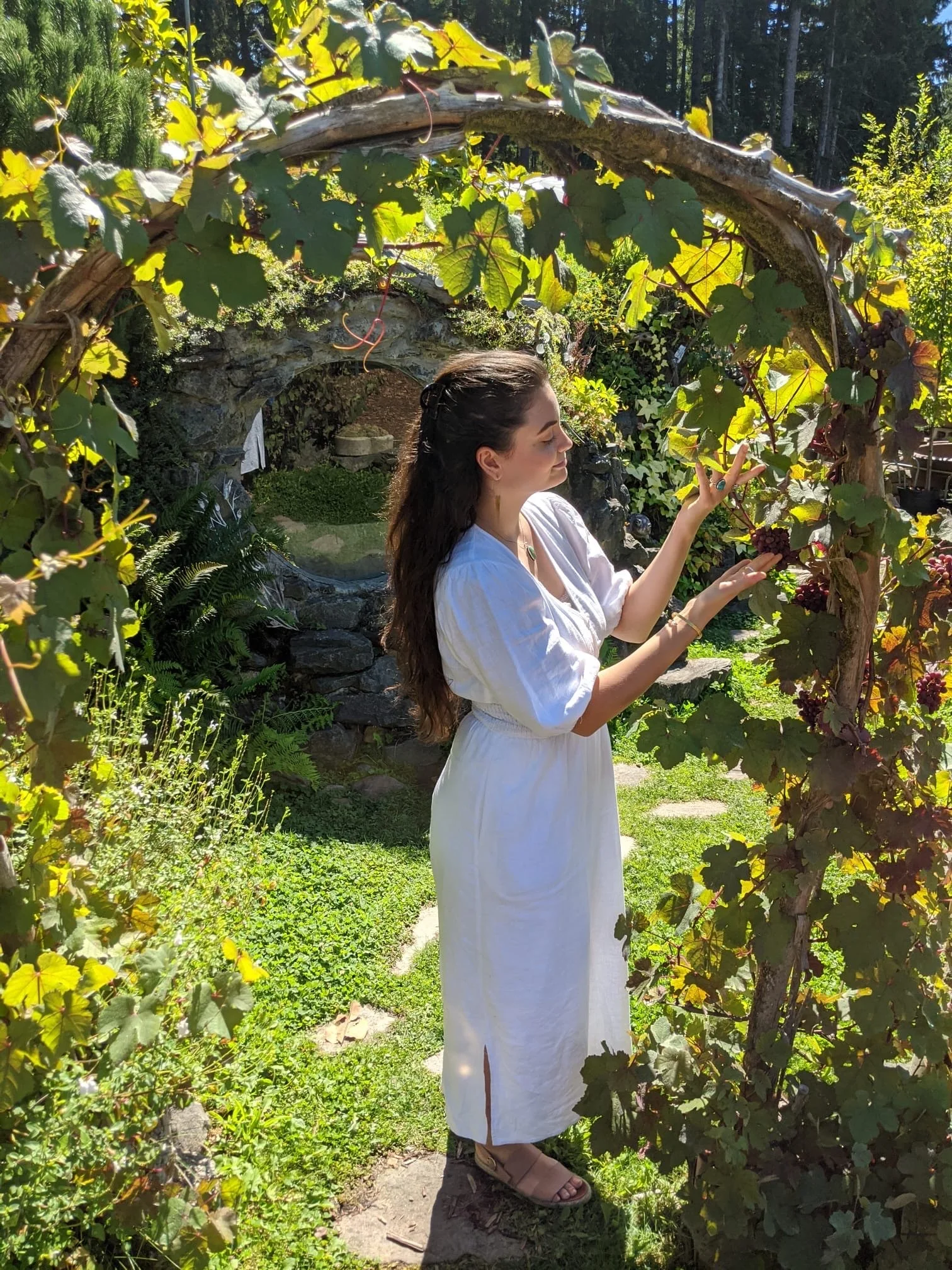A woman with long dark hair, wearing a white dress and sandals, standing in a garden under a grapevine arch with sunlight filtering through the leaves.