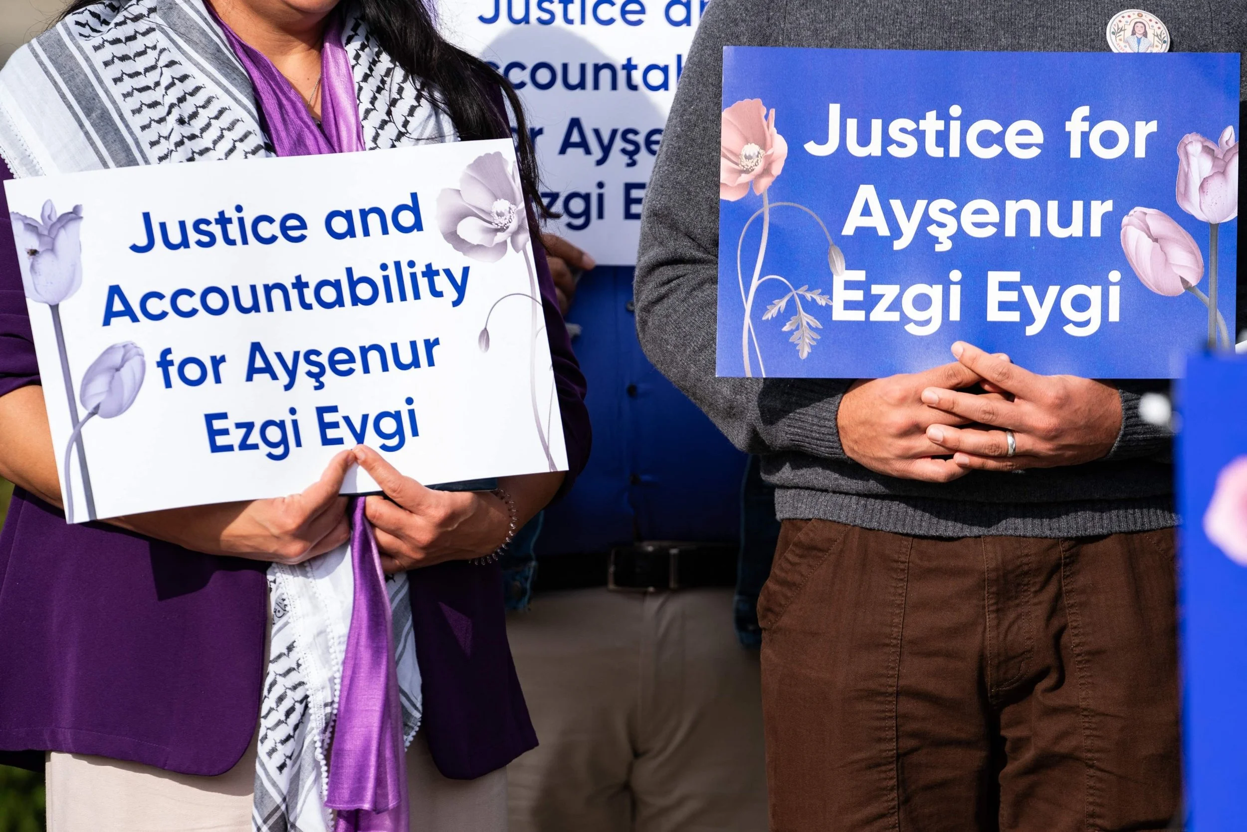 Two people holding signs that read "Justice for Ayşenur Ezgi Eydi." The signs are decorated with pink and purple flowers. The individuals are attending a public gathering or protest.