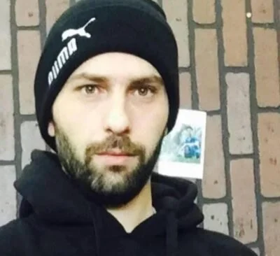 A man with a beard and mustache wearing a black Puma beanie and hoodie, standing in front of a brick wall.