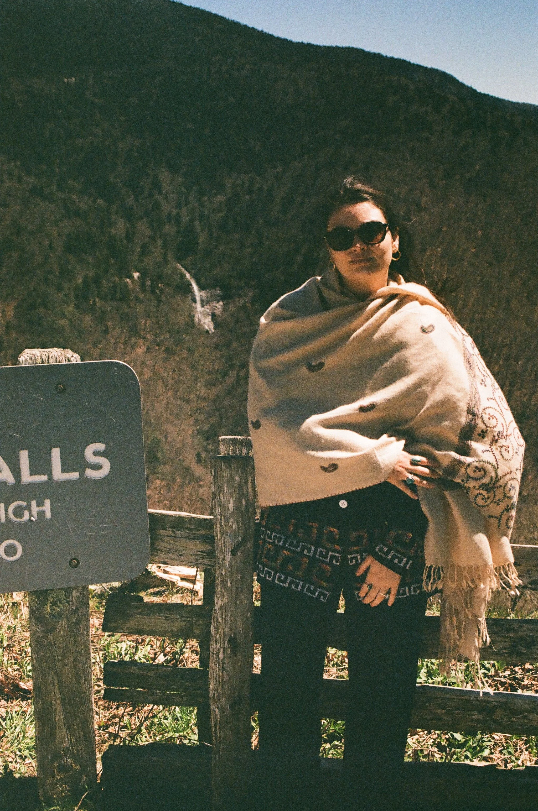 A woman wearing sunglasses and a beige shawl with embroidery, standing outdoors with a mountainous landscape in the background during daytime.