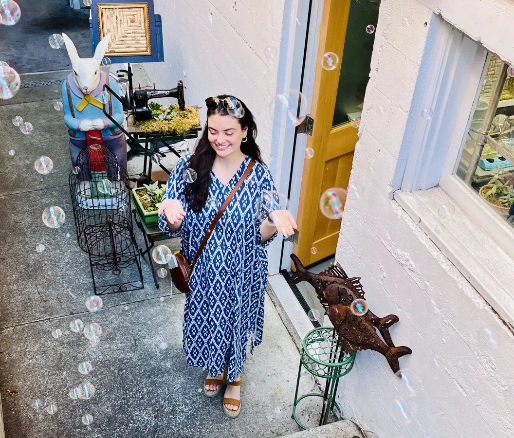 A woman in a blue and white dress standing outside near a white wall, surrounded by bubbles, with a smiling expression. Behind her, a rabbit statue wearing a vest and bow tie, a vintage sewing machine, and steps with decorative fish sculptures are vi