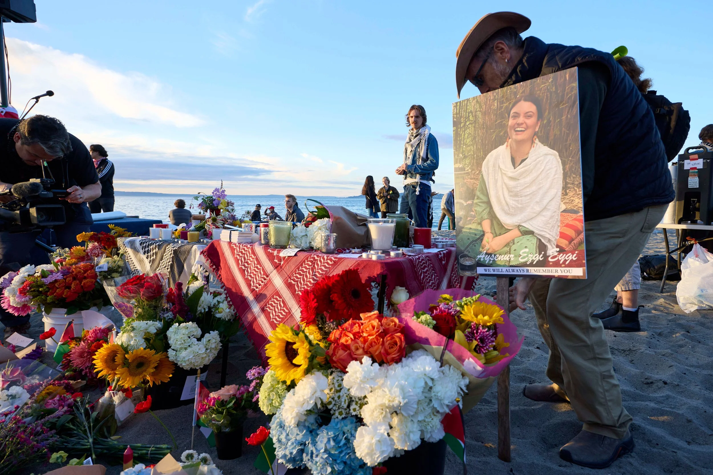 A memorial display on a beach featuring numerous colorful flower bouquets, candles, and a photo of a smiling woman with the name Ayşenur Ezgi Eygi and the message "We will always remember you". People are gathered around in the background, and the scene is set during the daytime with clear skies.
