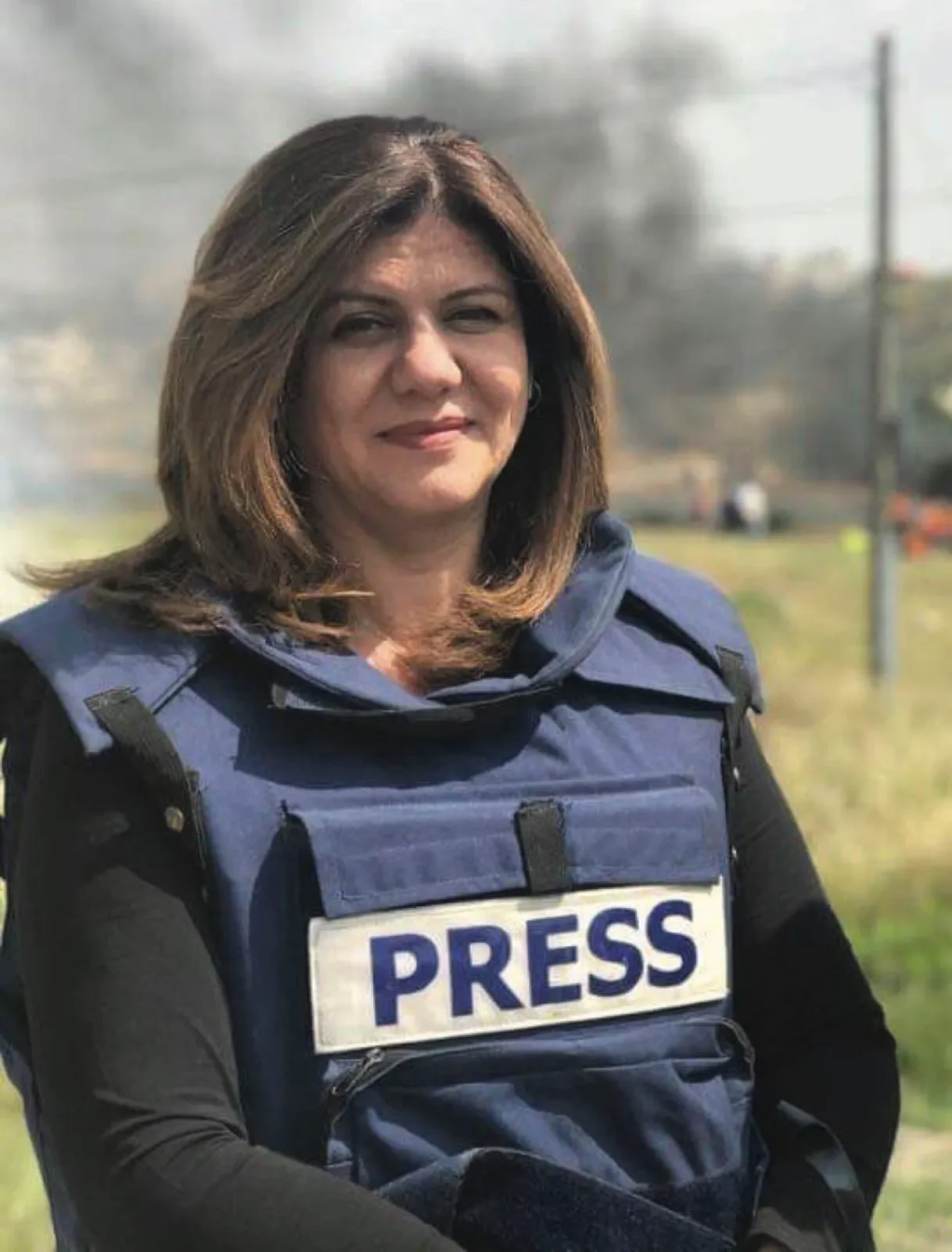 A woman with brown, shoulder-length hair, smiling, wearing a black long-sleeve shirt and a blue press vest, outside with a sunny sky and some trees in the background.