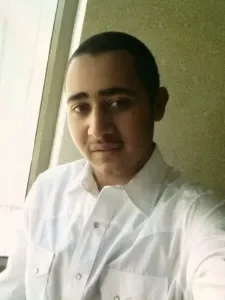 A young man with short dark hair, wearing a white button-up shirt, taking a selfie indoors near a window.