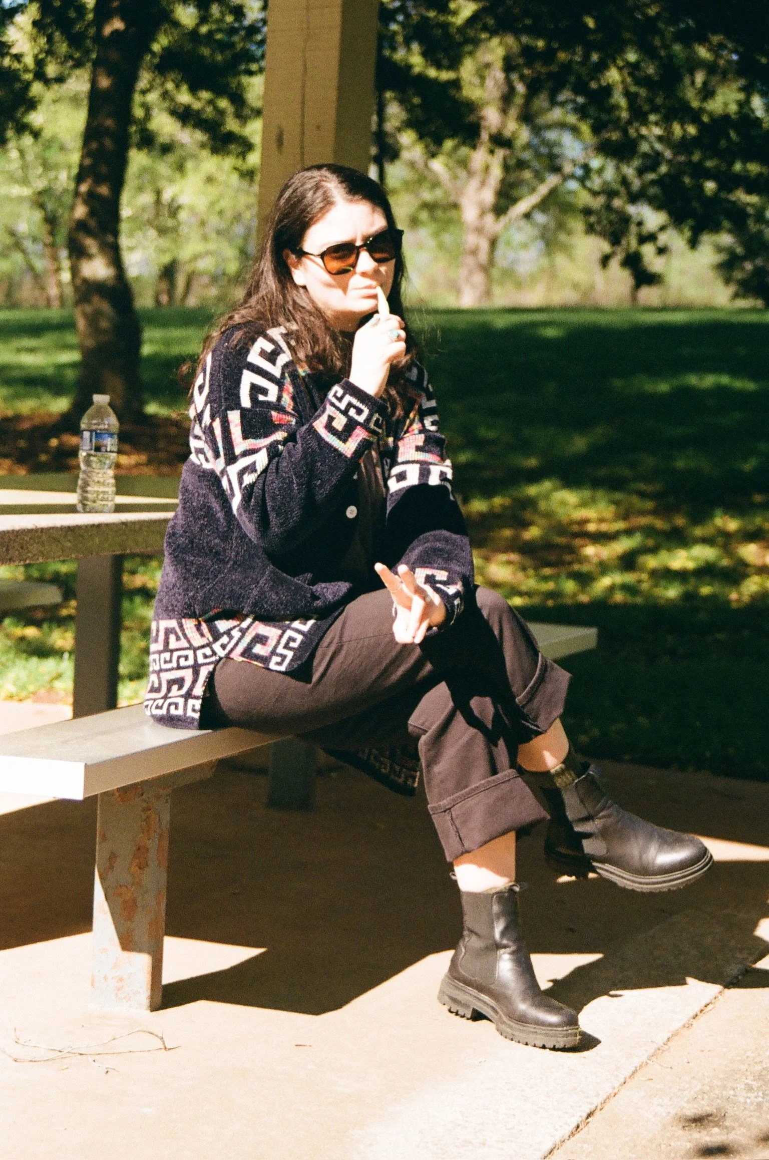 Woman with dark hair, sunglasses, and a patterned sweater sitting on a park bench, with a water bottle nearby, outdoors with trees in the background.