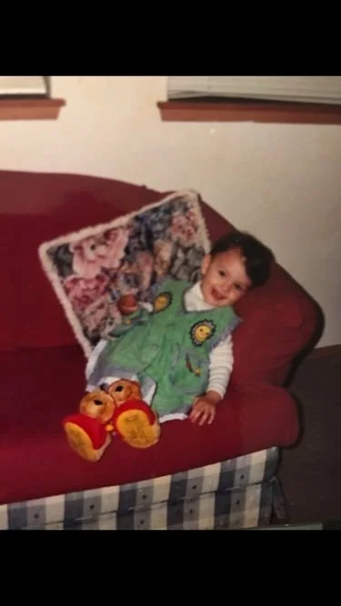 A young child sitting on a red couch with a colorful blanket, wearing a green, yellow, and white outfit, and red and yellow slippers, smiling at the camera.