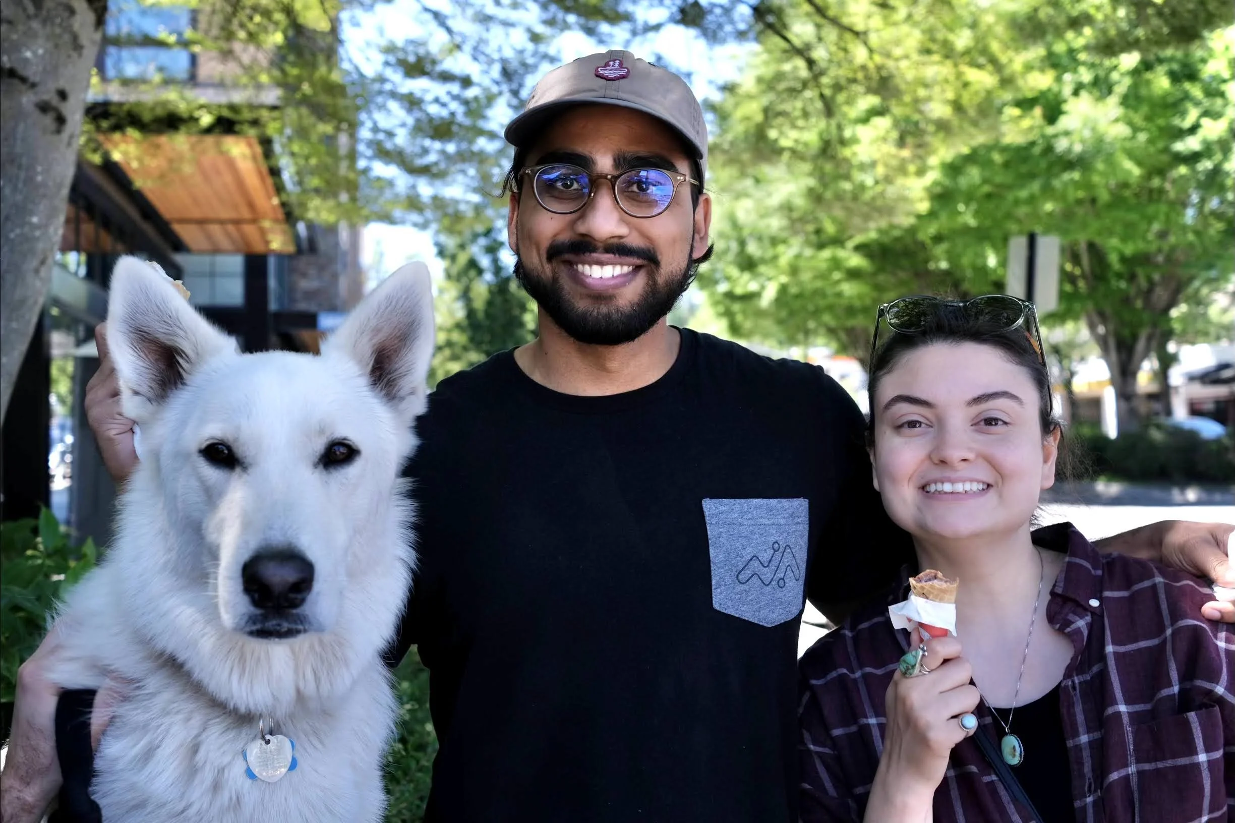 Two smiling people, a man wearing glasses and a hat, and a woman, stand outdoors with a white dog and a woman holding an ice cream cone. The background shows green trees and a building.