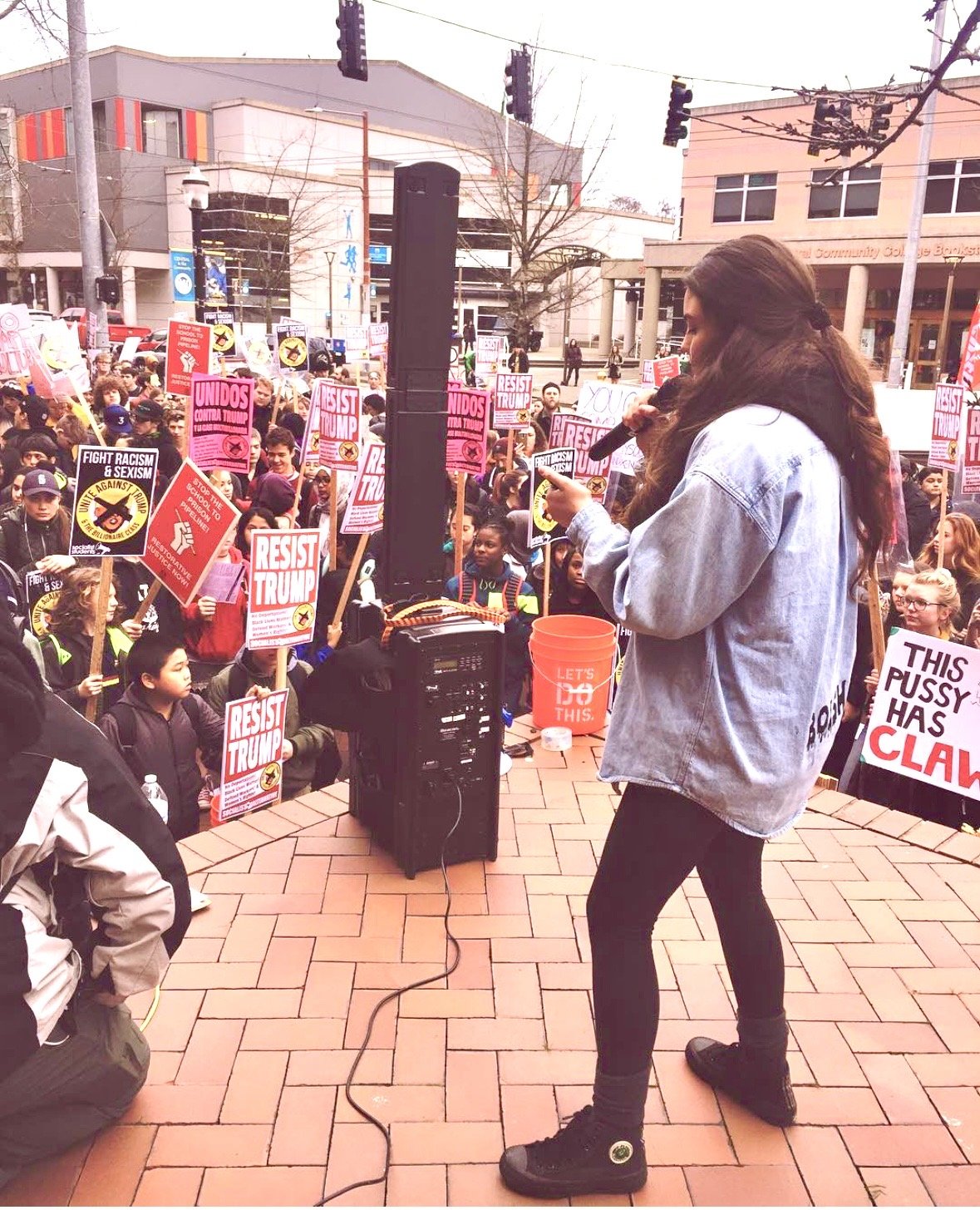 A woman standing on a brick-paved outdoor plaza speaking into a microphone at a protest or rally, with a crowd of young protesters holding signs expressing political messages, including signs that say 'RESIST TRUMP' and 'FIGHT RACISM & SEXISM'; buildings and traffic lights are visible in the background.