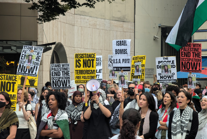 Protesters holding signs advocating for Palestine and ending occupation, with some signs reading 'Stand with Palestine! End the occupation now!' and 'Resistanc is not terrorism! Free Palestine!'. Others mention Israel bombs and support for Palestine, with a large crowd of people gathered in an outdoor space.