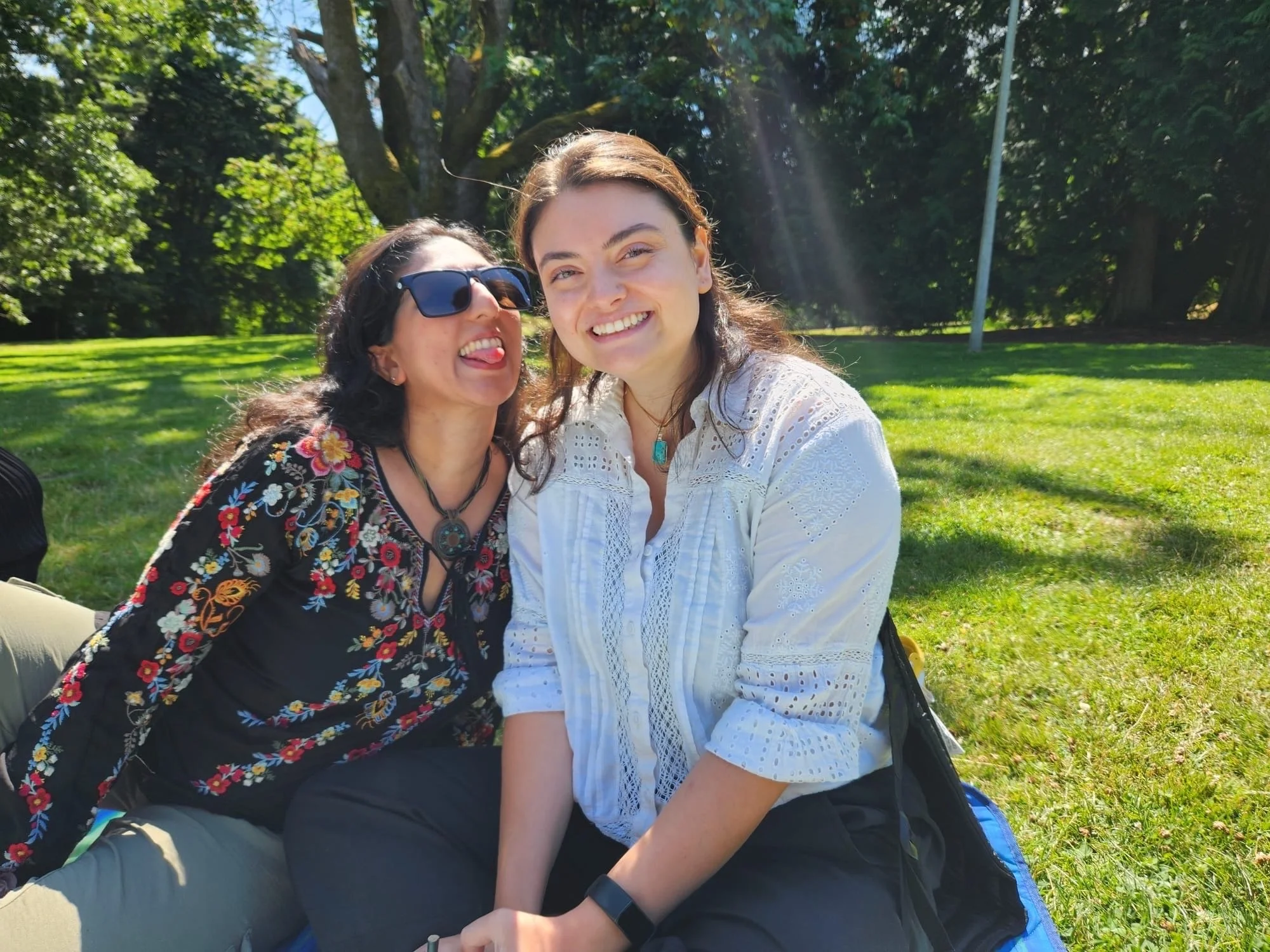 Two women sitting on grass in a park, smiling and enjoying sunny weather with trees and a blue sky in the background.
