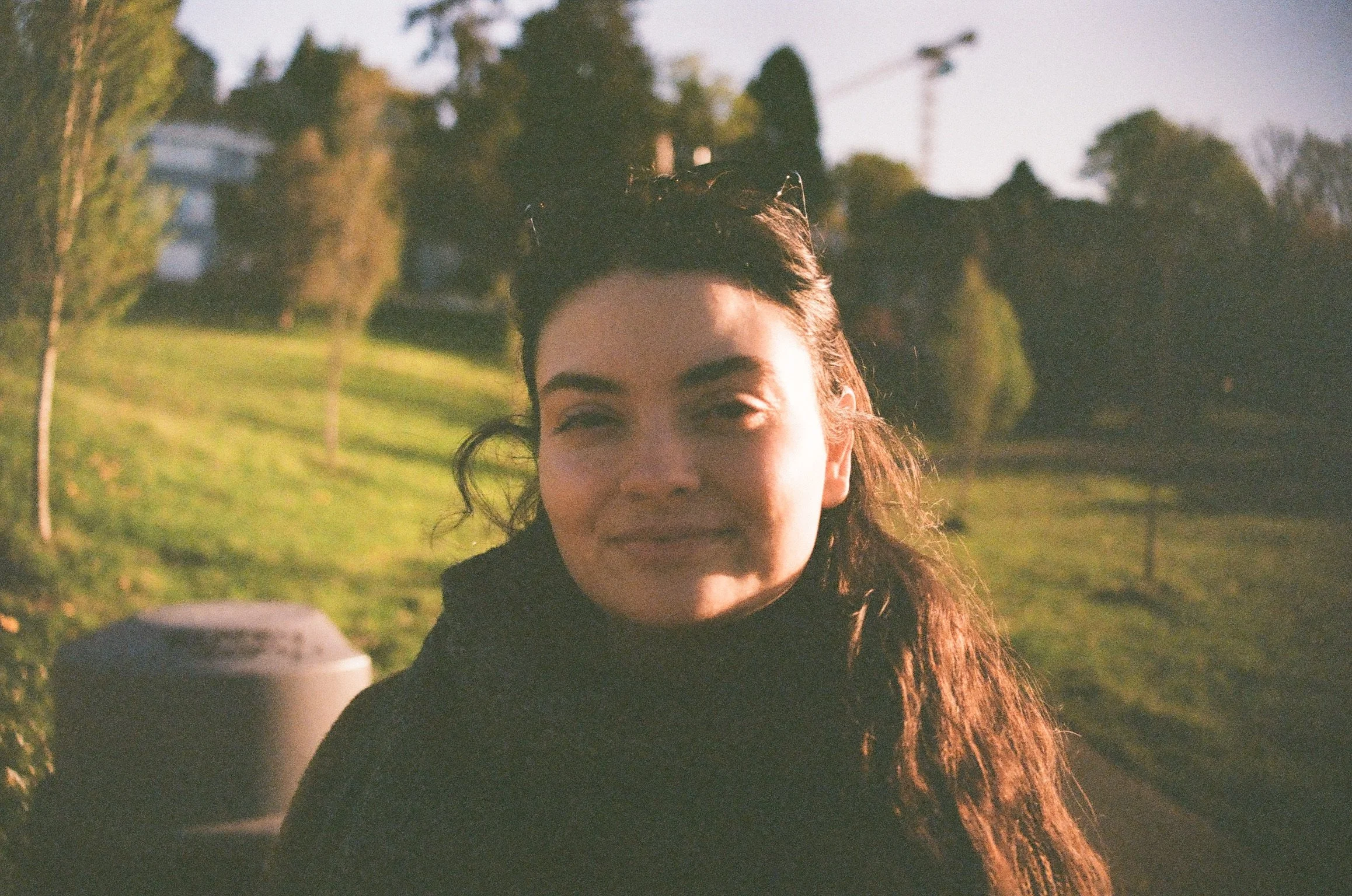 A woman smiling outdoors holding a coffee cup in a park with trees and grass in the background at sunset