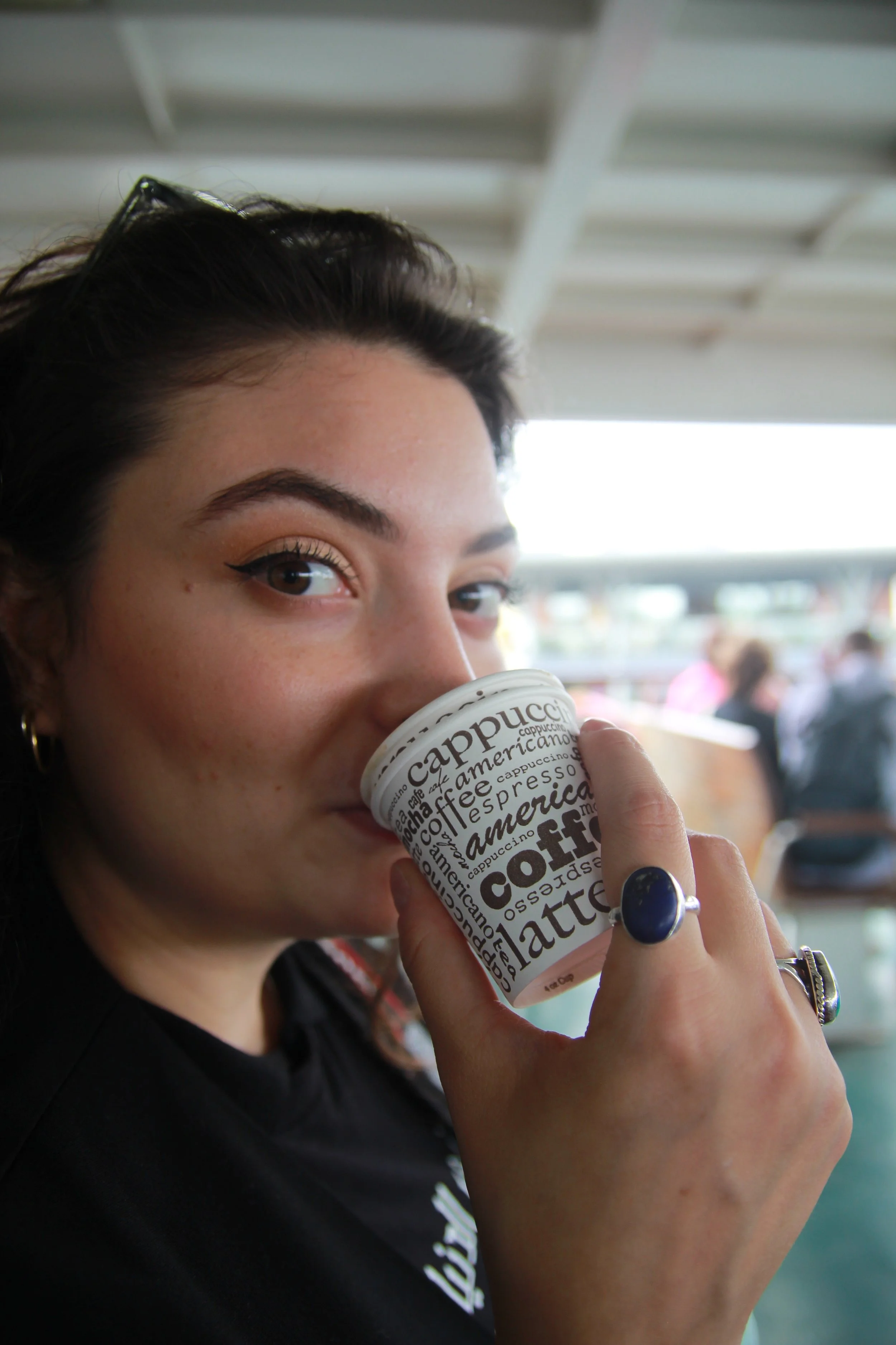 A woman with dark hair and eyebrows is holding a paper coffee cup near her face, with her eyes looking at the camera. The cup has black text with various coffee-related words. The background appears to be a public place with people and a roofed struc