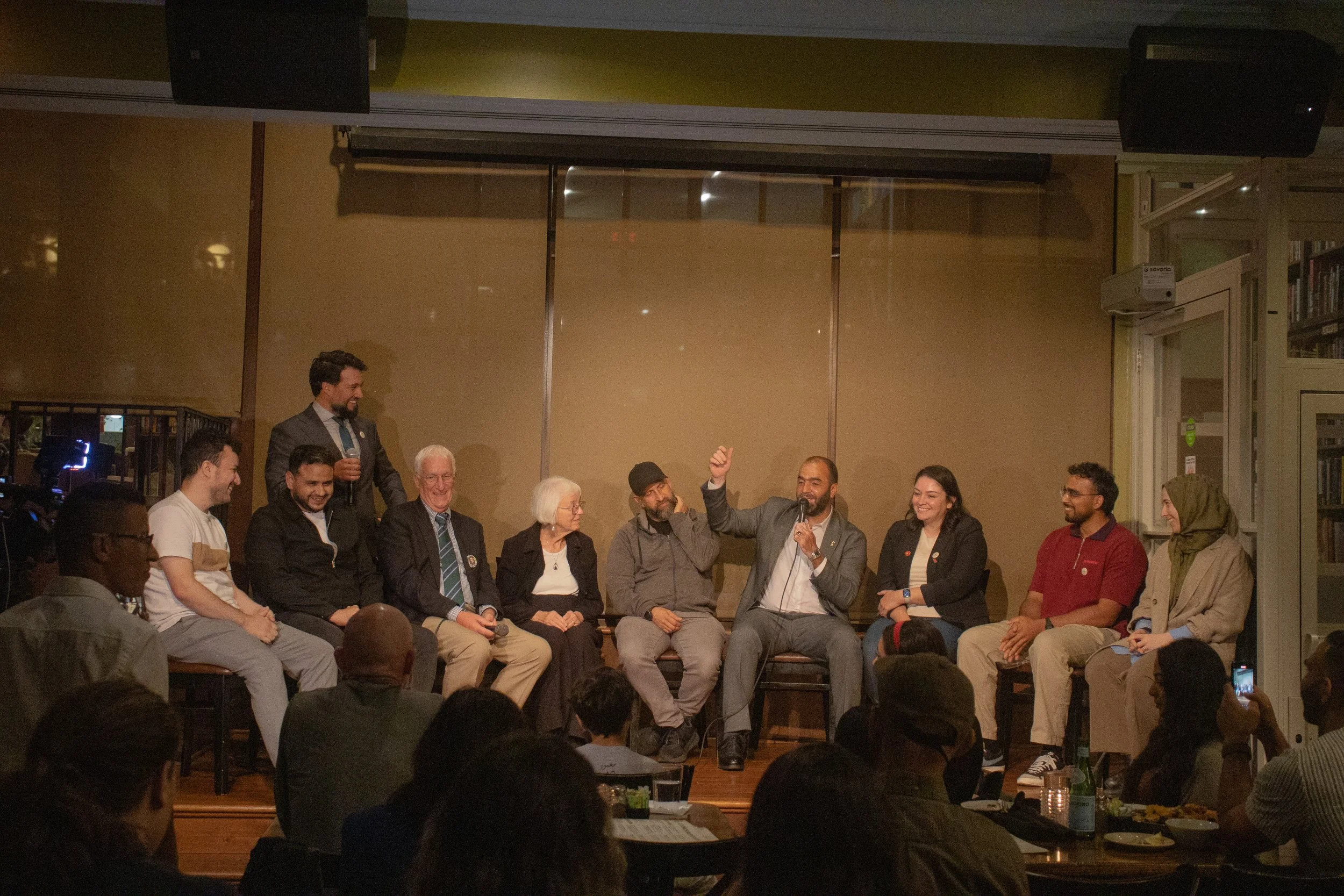 A group of diverse people sitting and standing in a room, engaged in a discussion or panel, with one man holding a microphone. The audience is visible in the foreground, and the setting appears to be an indoor event space or bookstore.