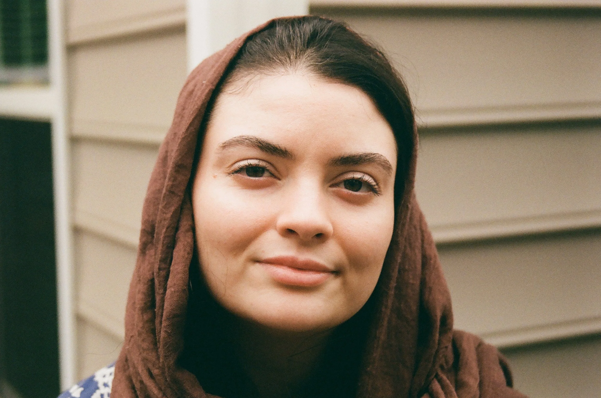 A close-up of Ayșenur Ezgi Eygi wearing a brown headscarf and a black top, smiling slightly against a blurred background of beige siding and some greenery.