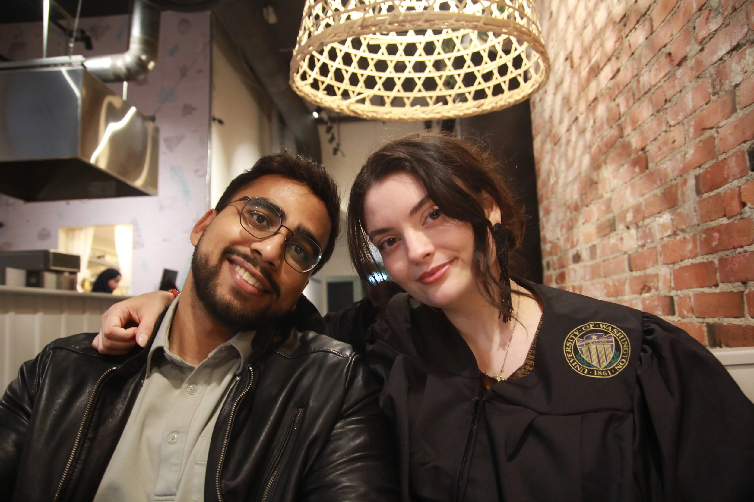 Two people smiling and posing for a photo in a restaurant, with a brick wall and a woven hanging lamp in the background. One of them is wearing glasses and a leather jacket, and the other is wearing a jacket with an emblem.