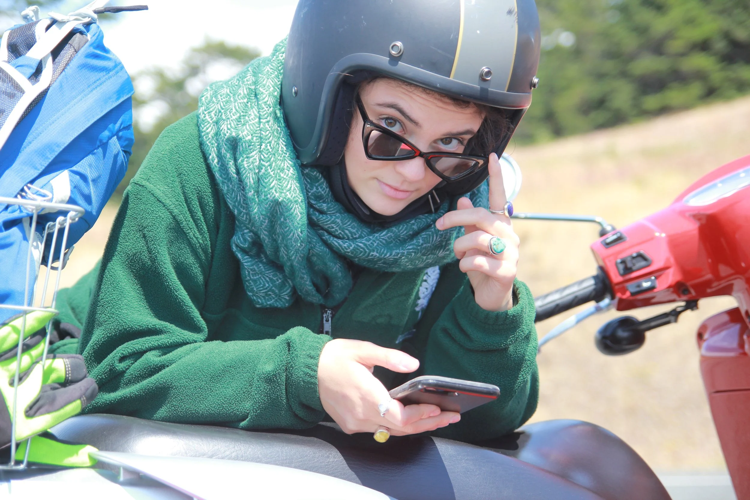Woman wearing a helmet and sunglasses, sitting on a motorcycle or scooter, adjusting her smartphone with a green jacket and scarf, outdoors on a sunny day.