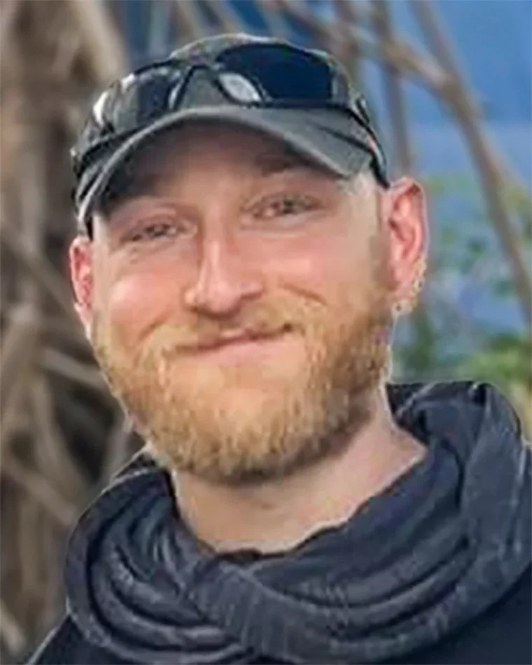 A man with a beard and mustache wearing a black cap and sunglasses, outdoors with trees and sky in the background.