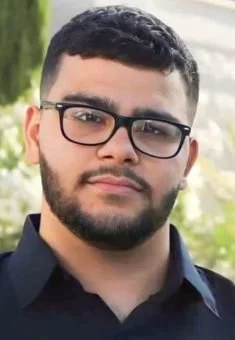 A young man with short dark hair, glasses, and a beard, wearing a dark collared shirt outdoors.