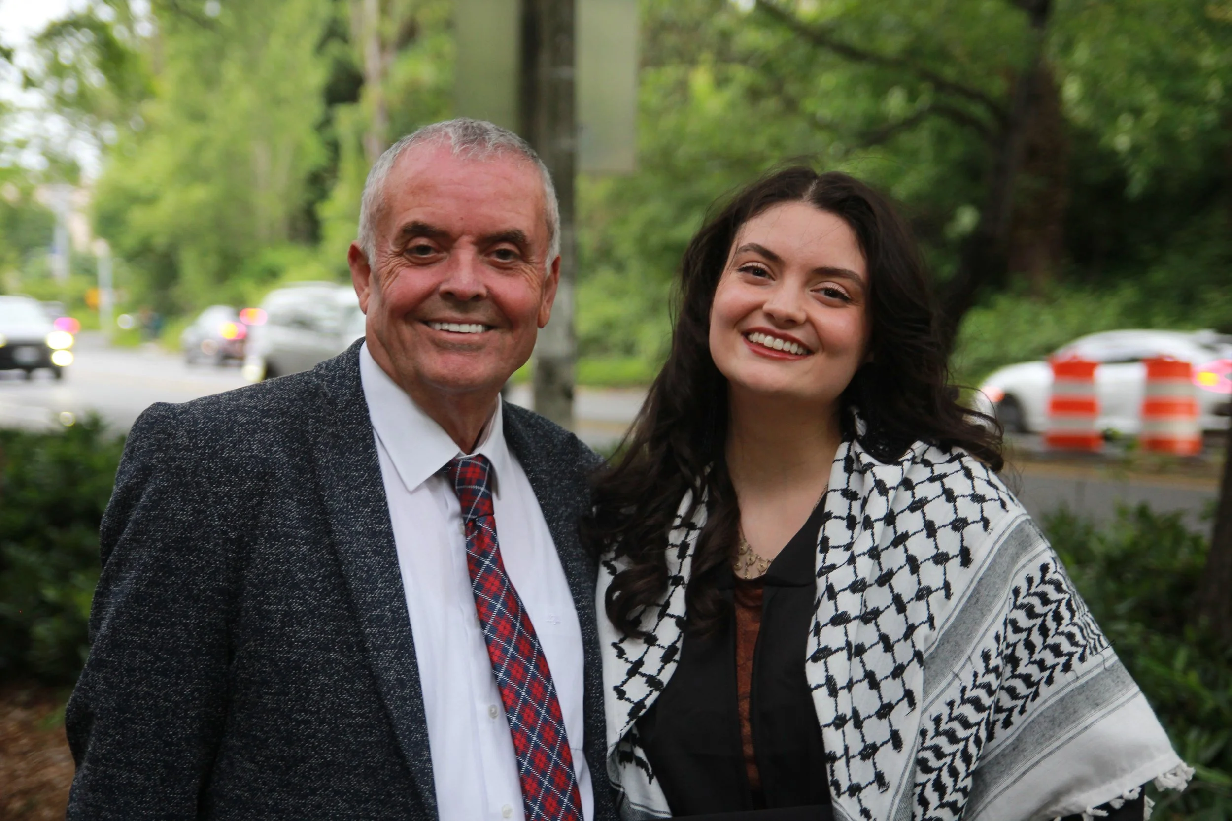 A man and a woman smiling outdoors in a park with trees, cars, and orange traffic barrels in the background.