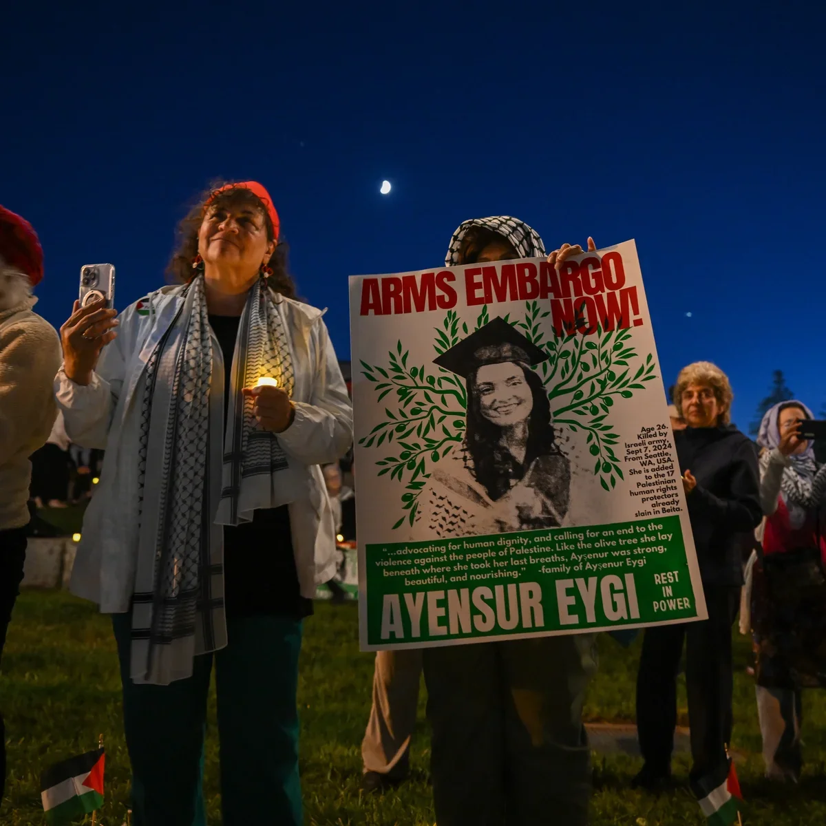 A woman holding a sign with a photo of a young woman, the words 'Arms Embargo Now!' in red, and information about Aysenur Eggi, who was killed in Israel. Others stand around during a nighttime protest under a dark blue sky with a crescent moon visible.