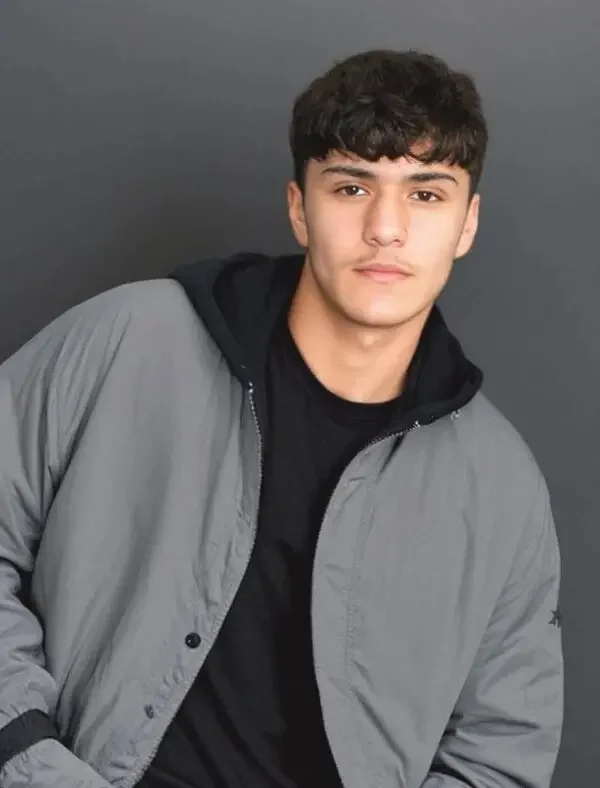 A young man with dark curly hair, light skin, wearing a black shirt and a gray jacket, standing against a plain dark background.