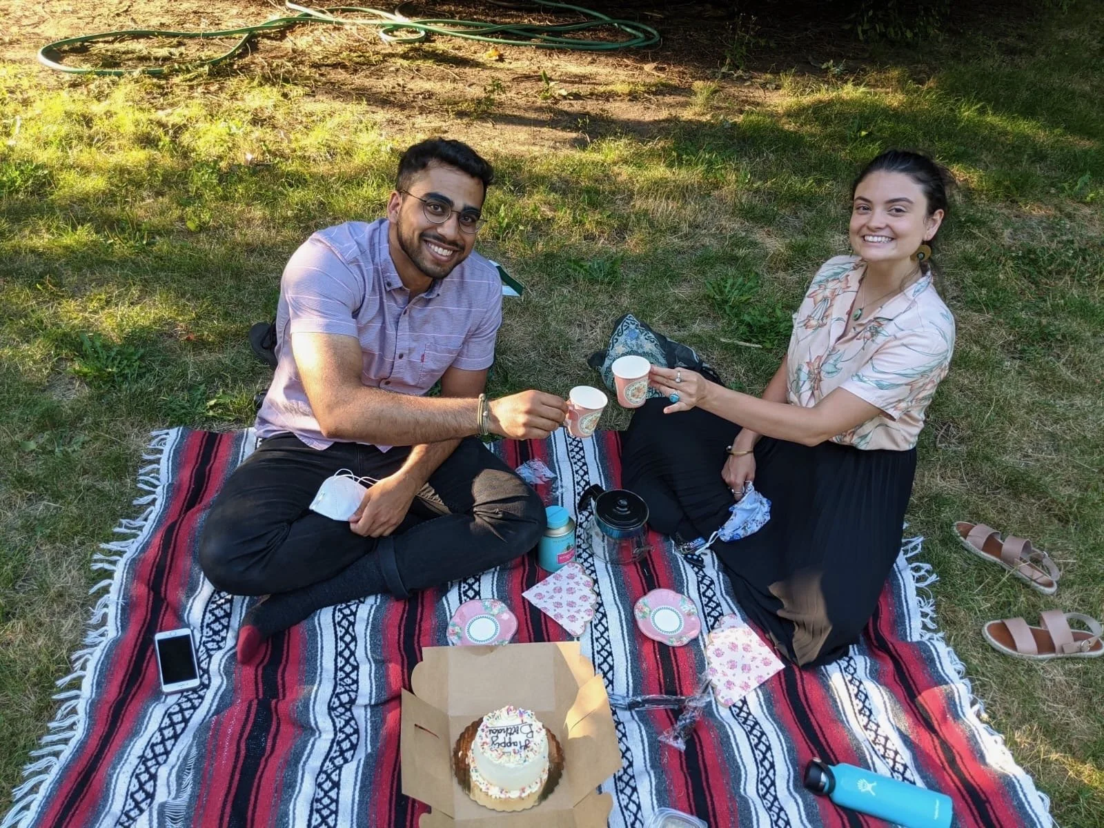 Two young adults having a picnic outdoors, seated on a colorful rug with a birthday cake, gift, and cups, smiling and clinking cups.