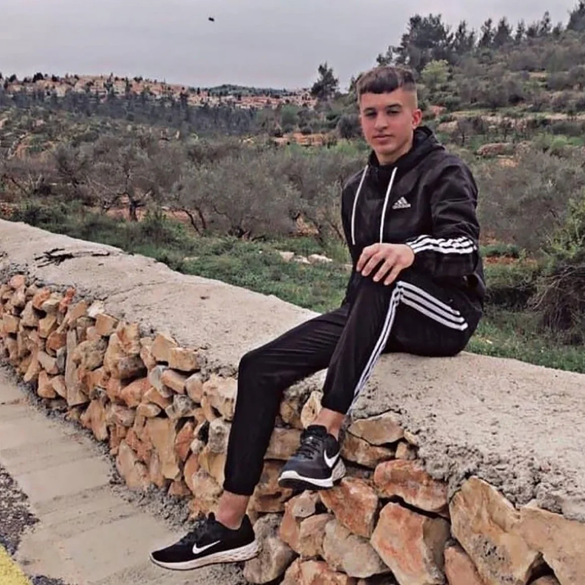 A teenage boy in a black Adidas tracksuit with white stripes sits on a stone wall in a rural landscape with trees and hills in the background.