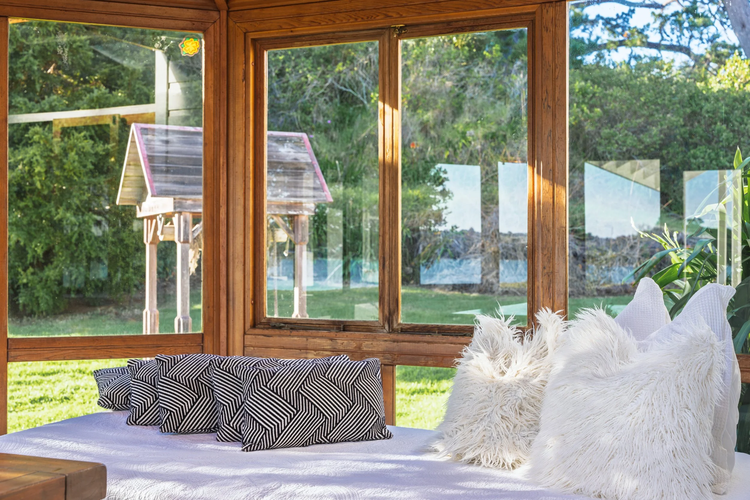 A cozy sunroom with a window seat decorated with patterned black and white pillows, fluffy white pillows, and surrounded by large glass windows showing green trees and a small outdoor structure with a pink roof.