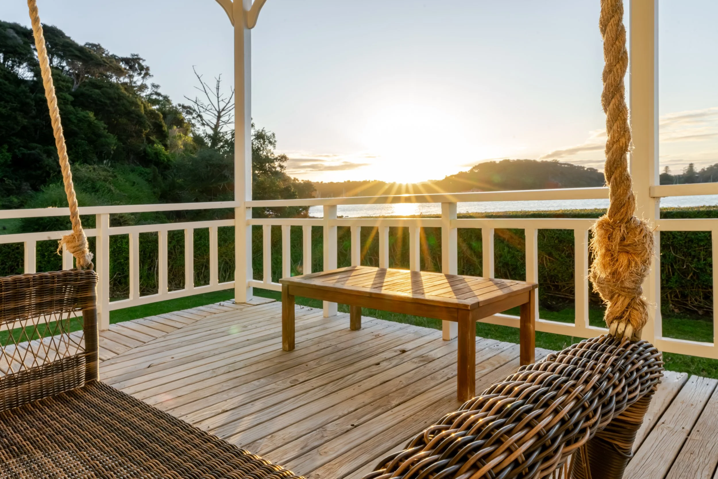 Sunset view from a wooden balcony with wicker furniture and a wooden bench, overlooking greenery and a lake.