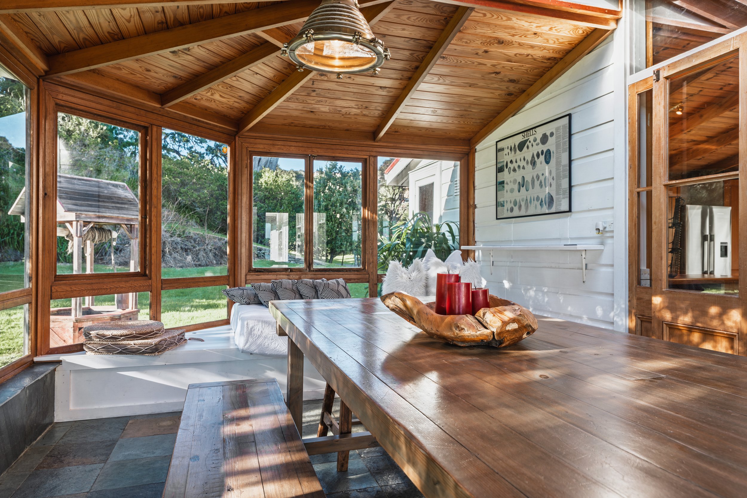 A sunlit enclosed porch with wooden ceiling and trim, large windows showing greenery outside, a wooden dining table with a wooden tray holding red candles, and a seating area with white cushions and throw pillows, adjacent to a white exterior wall wi