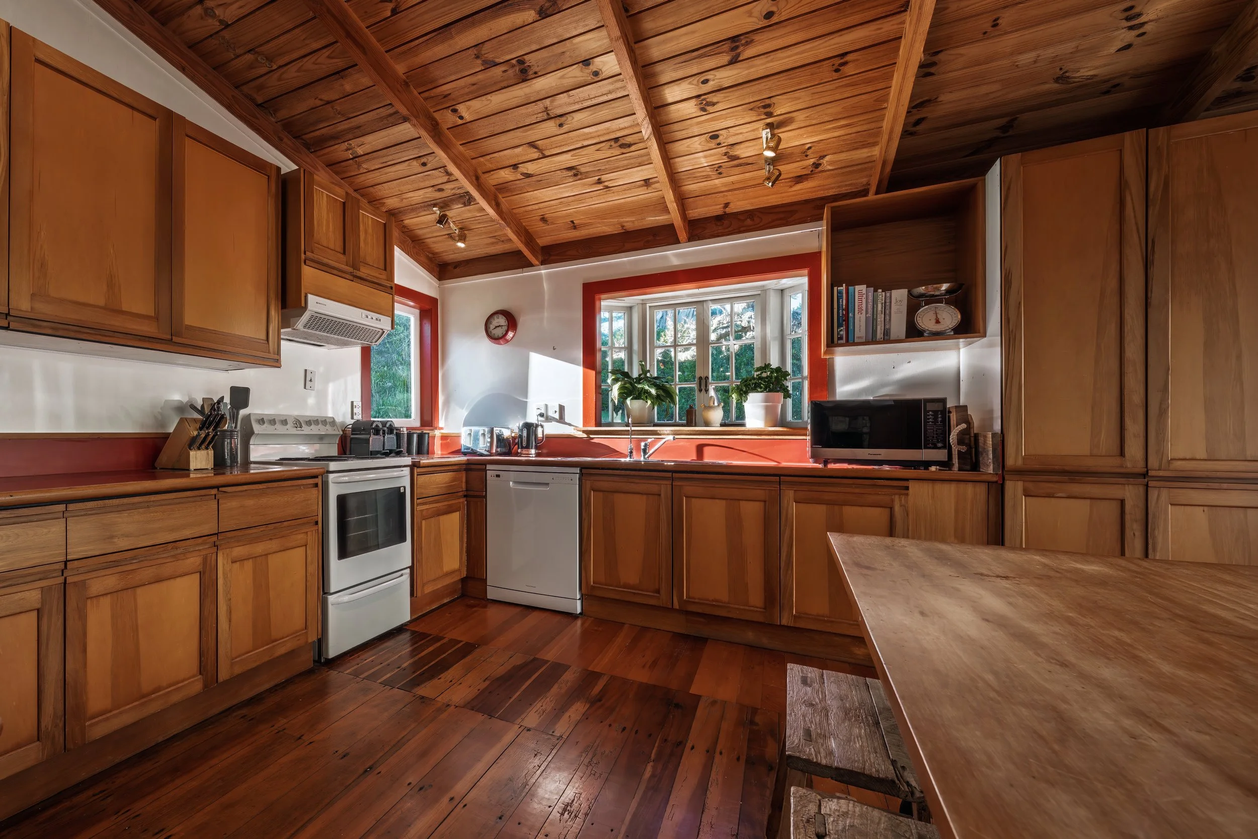 A cozy kitchen with wooden cabinets, a white stove, a dishwasher, and a microwave. Sunlight streams through a window above the sink, which has potted plants and kitchen appliances on the counter. The ceiling is wooden with exposed beams.