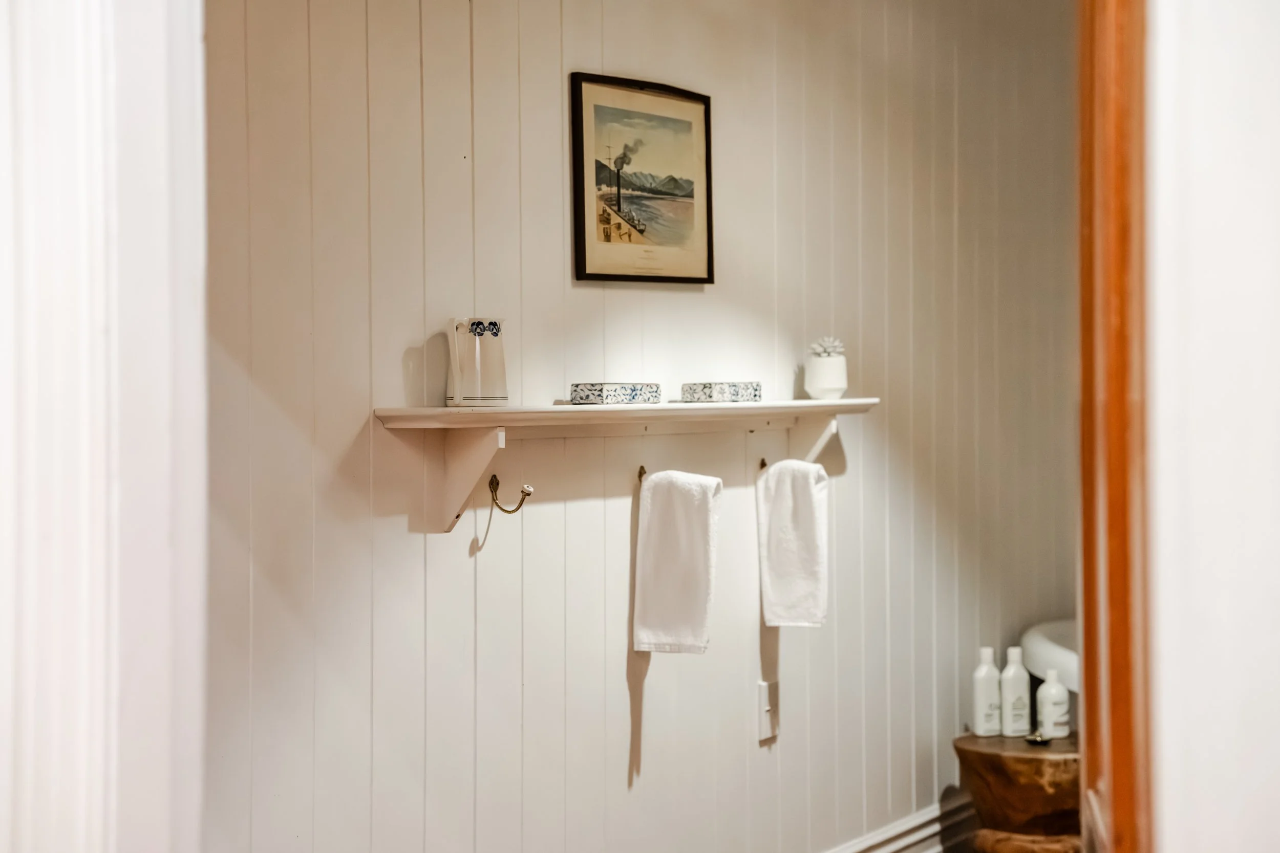 A bathroom with horizontal white wood panel walls, a framed painting of a landscape, a white shelf with two white towels hanging below, a small potted plant, and bottles of toiletries on a small wooden table.