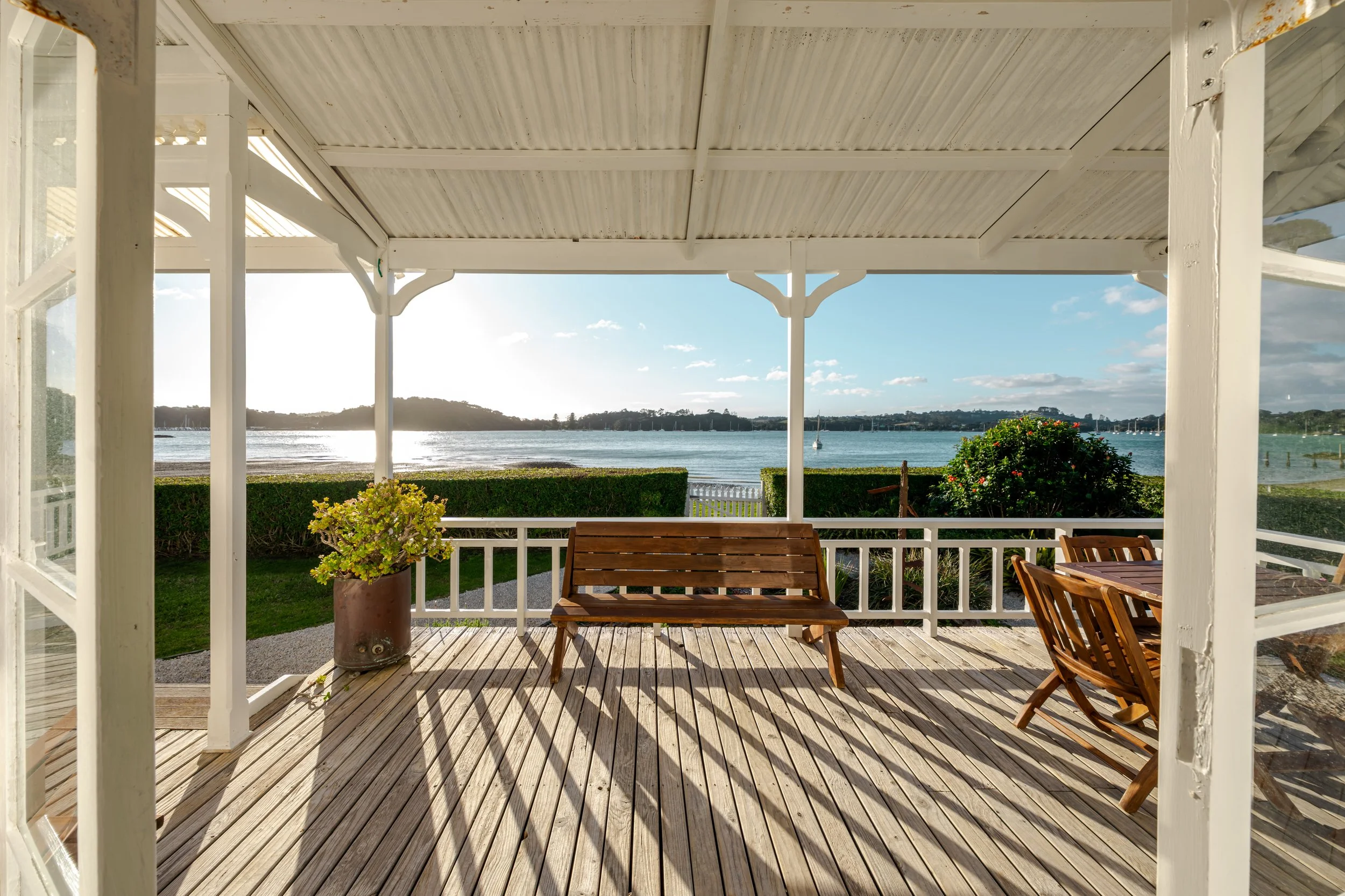 View from a screened porch overlooking a lake with boats, a hedge, and a cloudy sky, with a wooden bench and table on the porch.