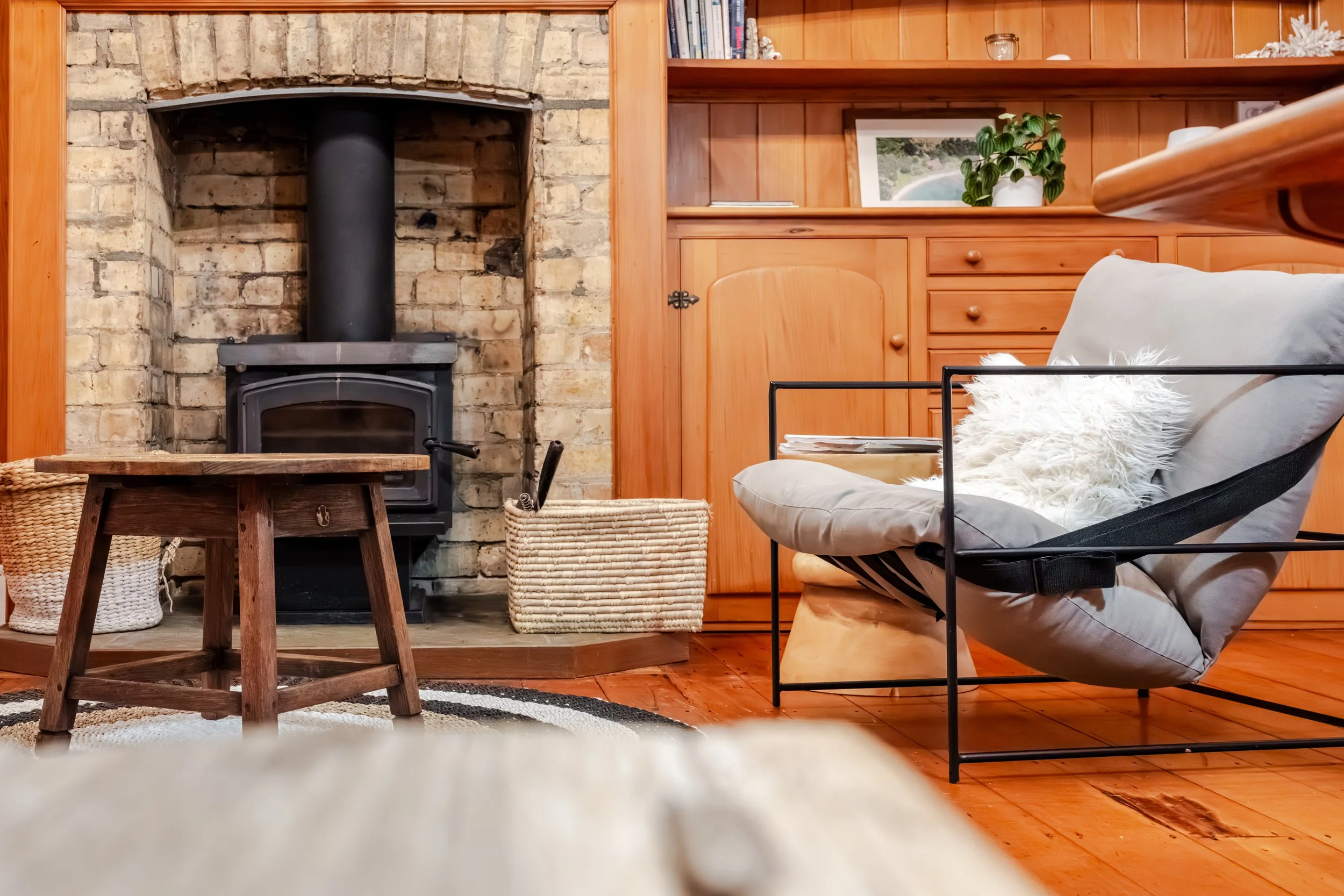 Living room with brick fireplace and wood-paneled wall, black wood stove, wicker baskets, wooden stool, light-colored armchair with white fluffy pillow, and wooden shelving with framed photo and plants.