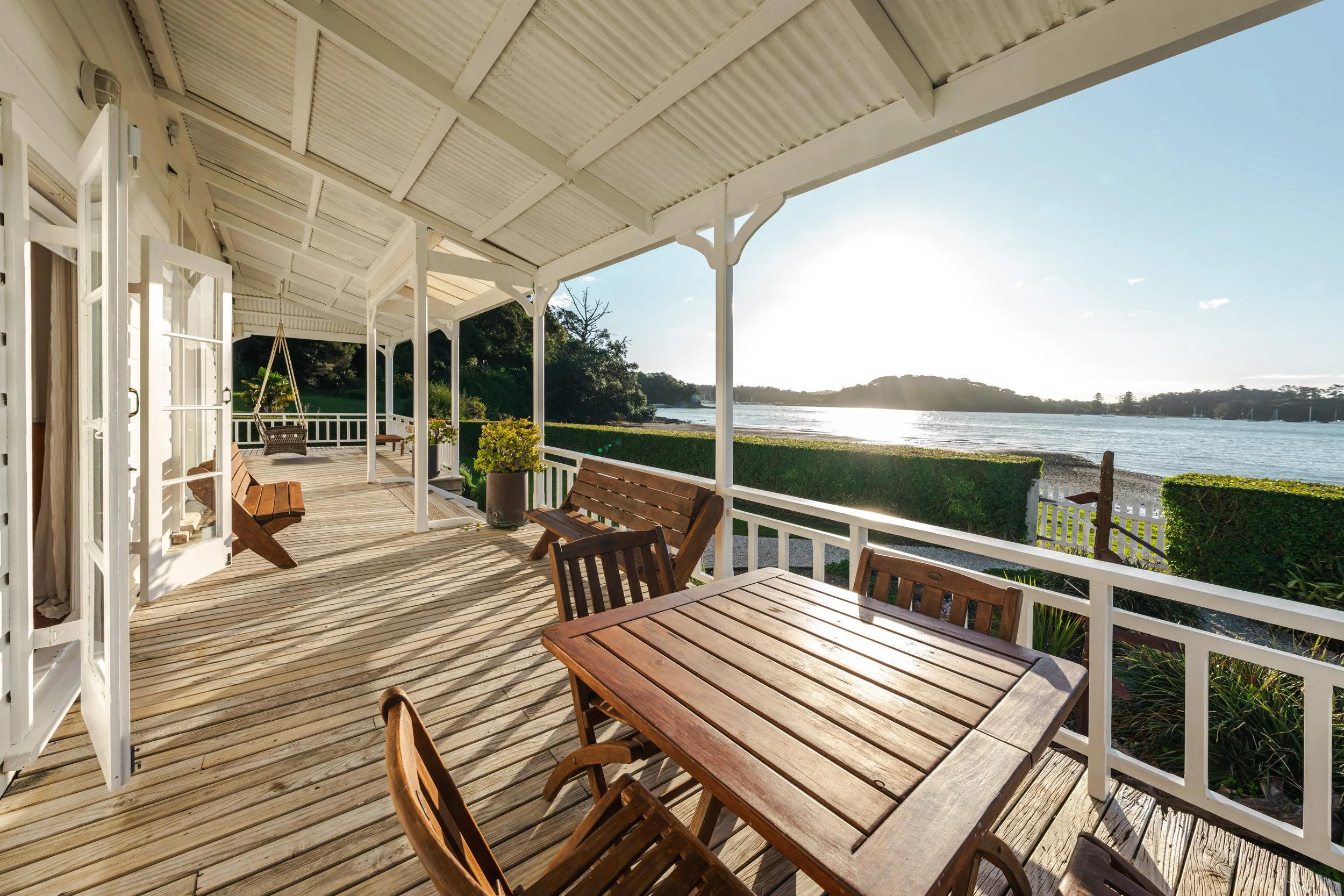 A wooden porch overlooking a body of water with a table and chairs, potted plants, and a swing, with sunlight reflecting off the water in the background.