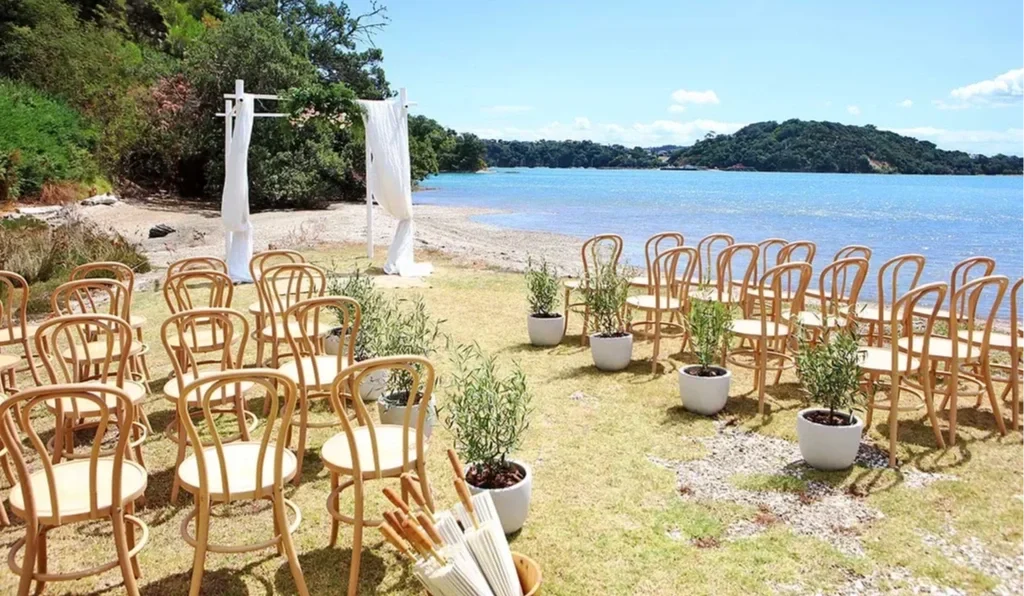 Outdoor wedding ceremony setup on a grassy lakeside with rows of chairs, potted plants, and a white fabric-draped arch, with water and hilly landscape in the background.