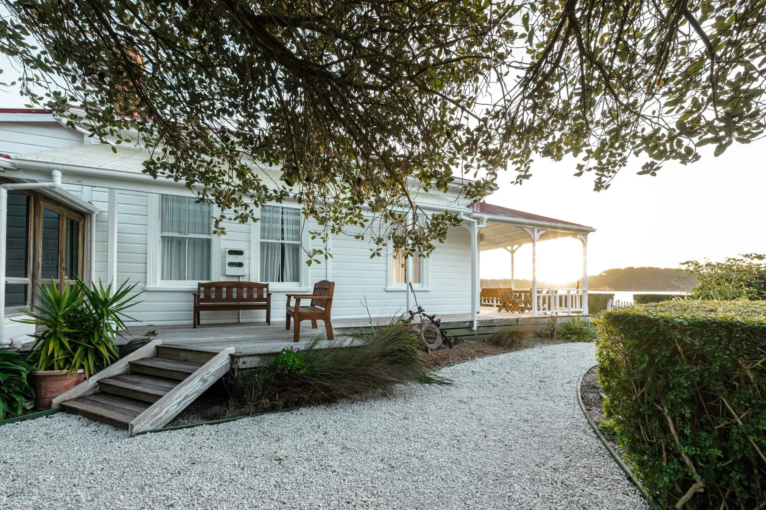 A white house with a wooden porch, outdoor seating, a gravel pathway, and surrounding green bushes and trees at sunset.