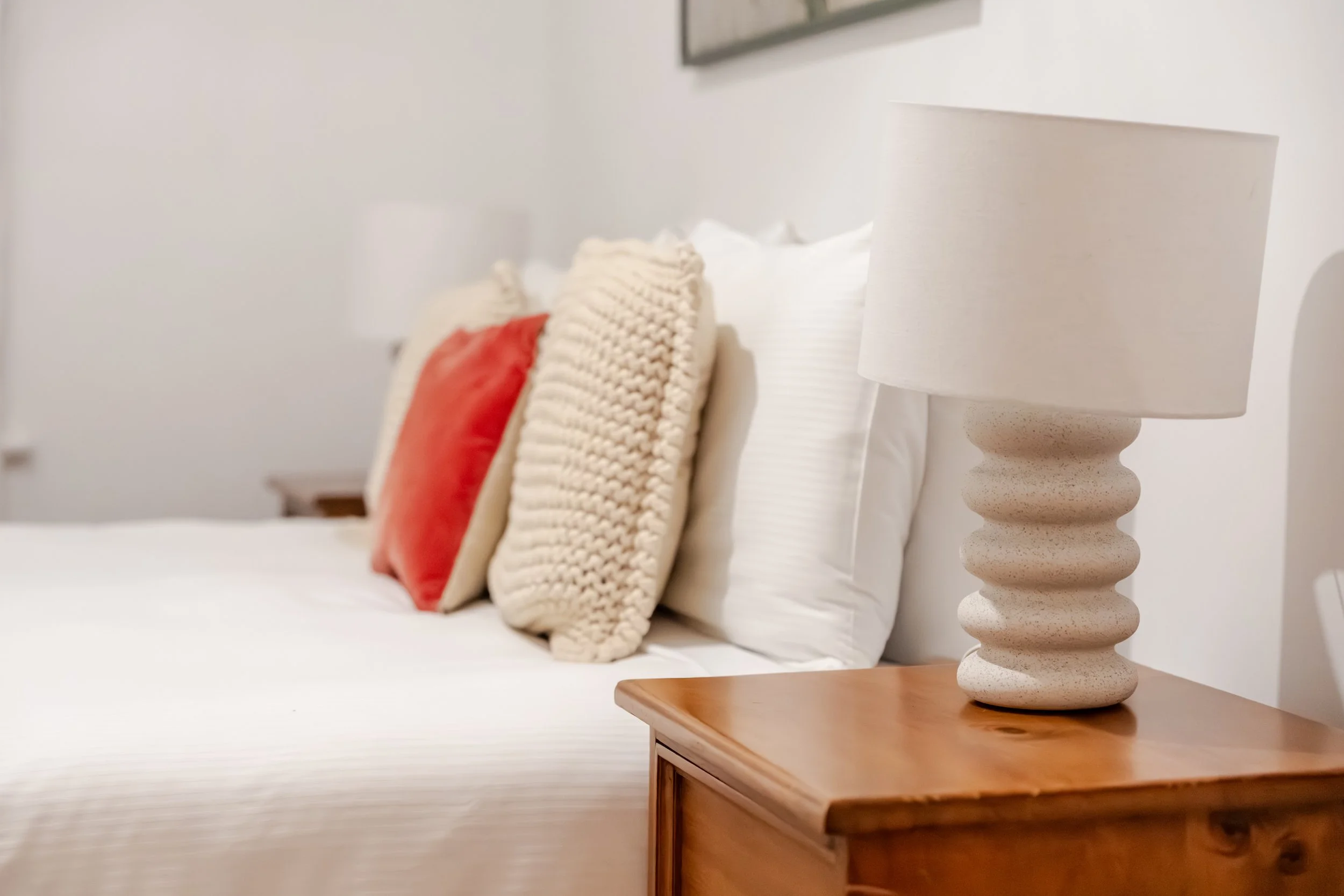 Close-up of a bedroom nightstand with a white textured lamp, a wooden nightstand edge, and decorative pillows on a bed, including a red pillow and cream-colored knitted pillow.