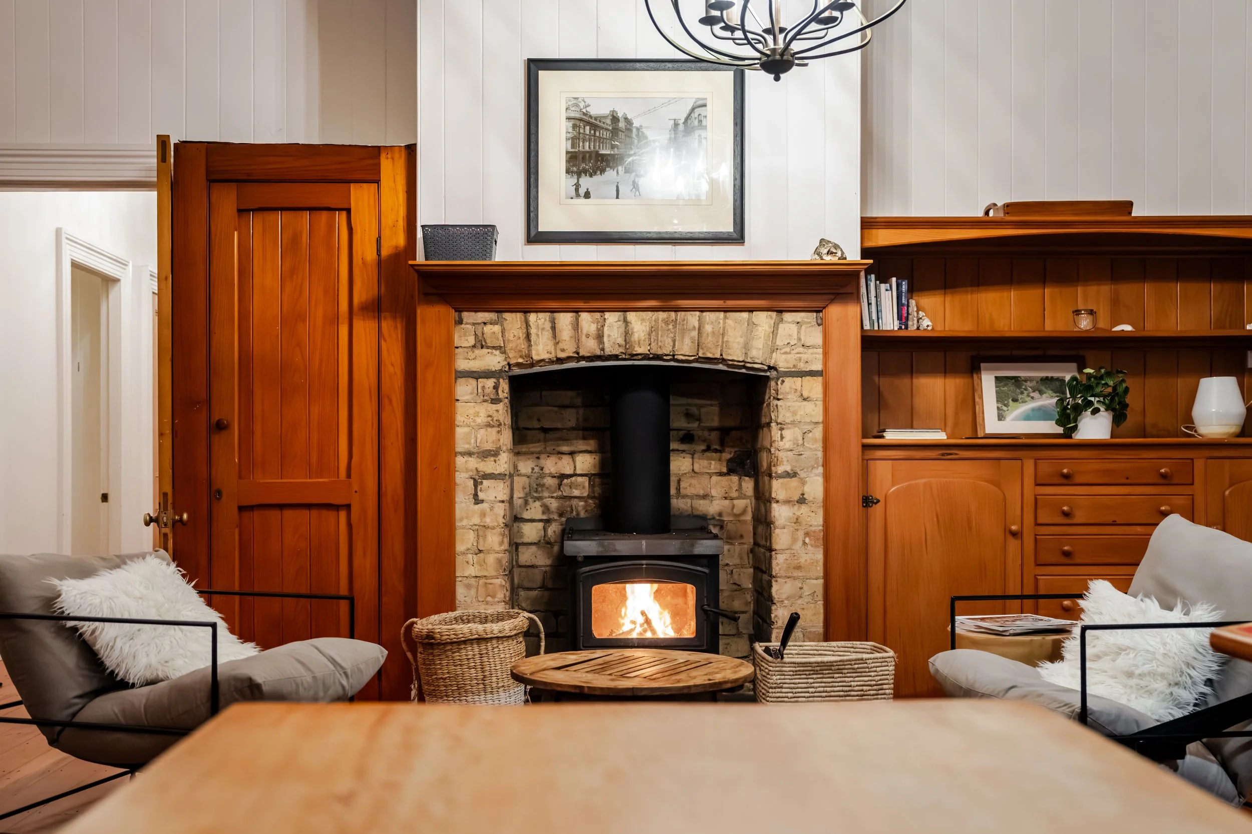 Living room with a brick fireplace, wood paneling, and a wood mantel. A burning fire in a wood stove inside the fireplace. Two armchairs with cushions and furry pillows, a round wooden table, woven baskets. A black-and-white framed photo hangs above 