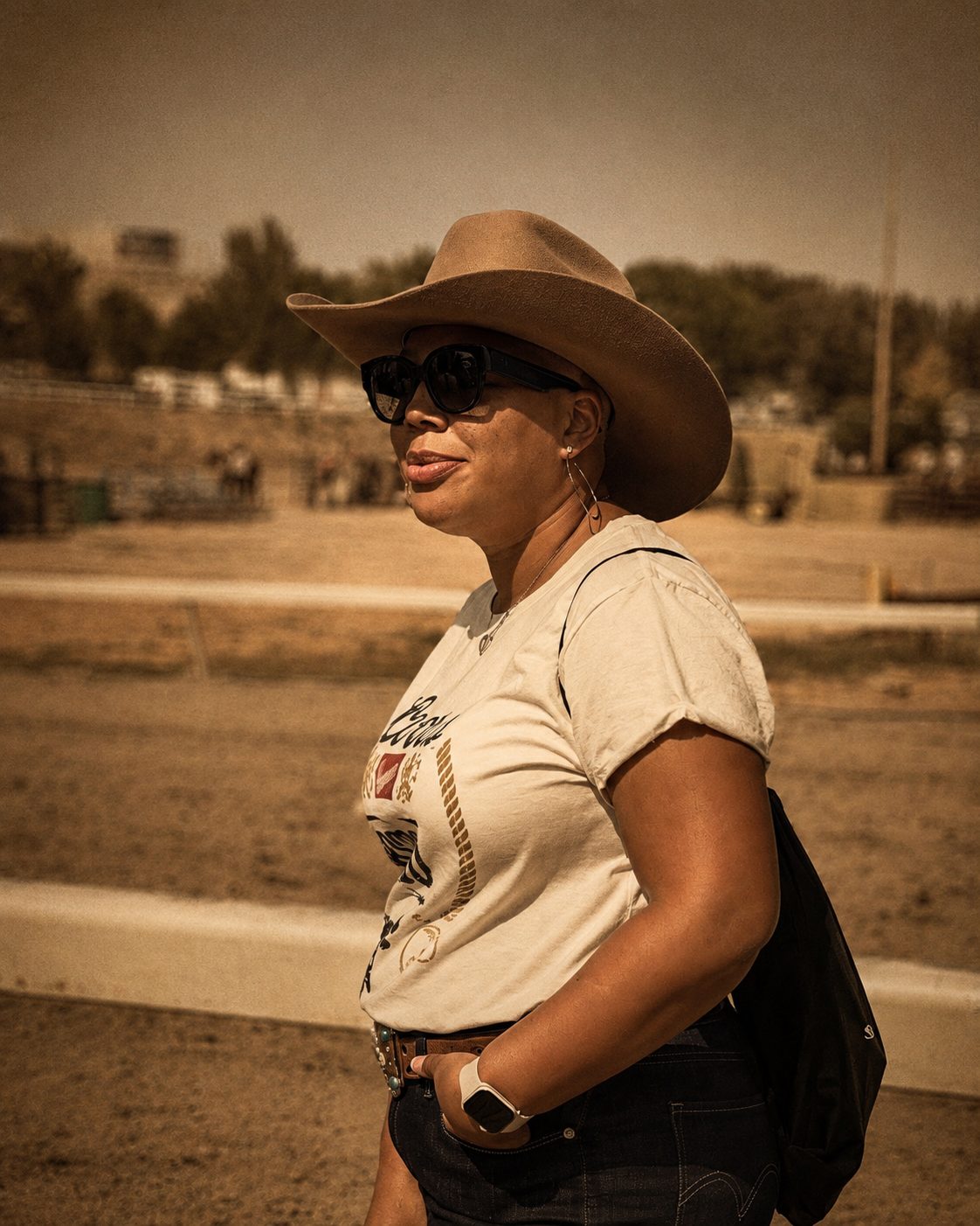 A woman wearing a tan cowboy hat, large black sunglasses, earrings, a graphic t-shirt, black jeans, a watch, and carrying a black bag, standing outdoors on a dirt path with trees and fencing in the background.
