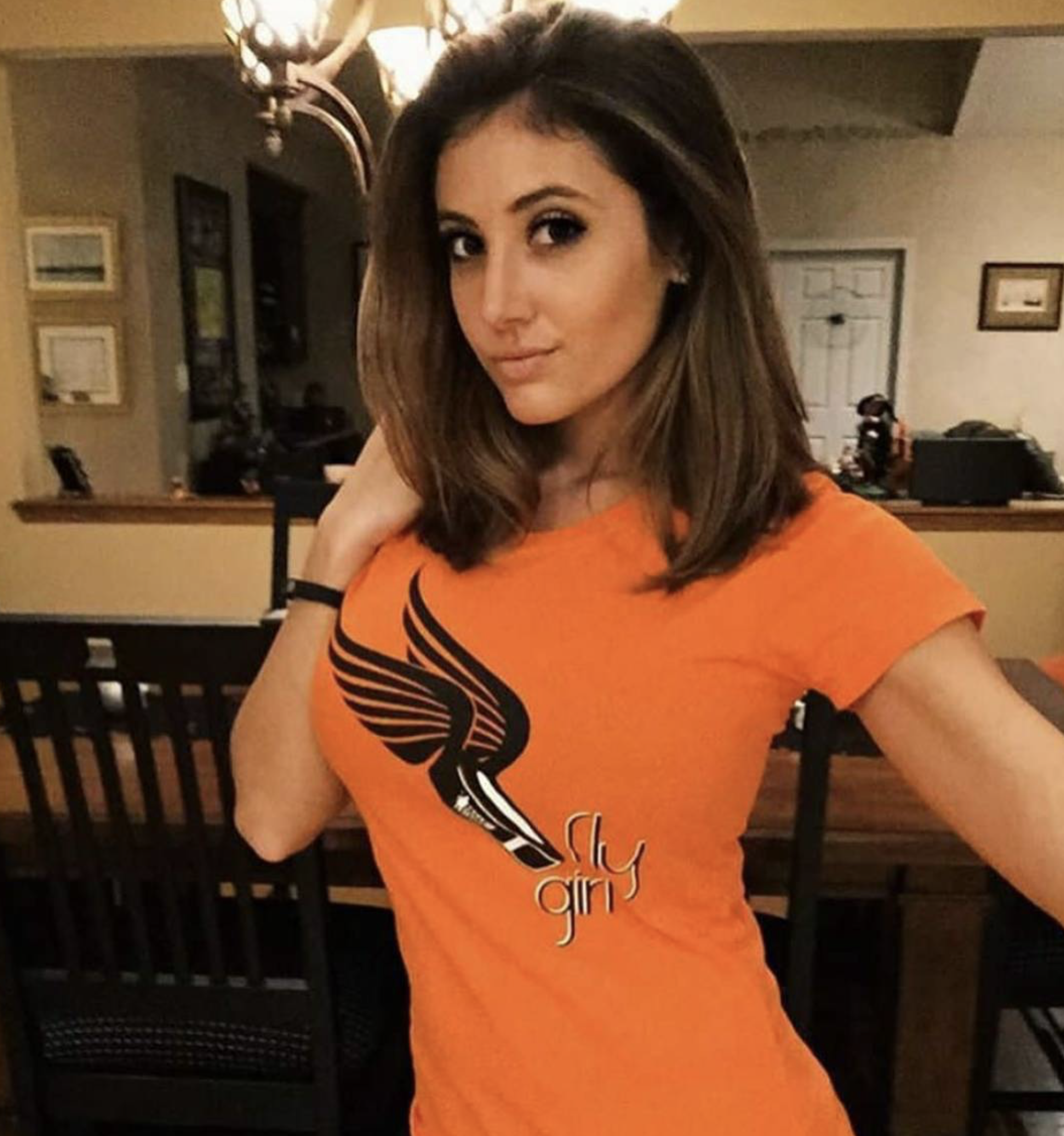 A young woman with shoulder-length brown hair and light skin posing indoors. She is wearing an orange T-shirt with a black winged emblem and the words 'fly girl.' She has a slight smile, one hand touching her hair, and is standing in front of a dinin